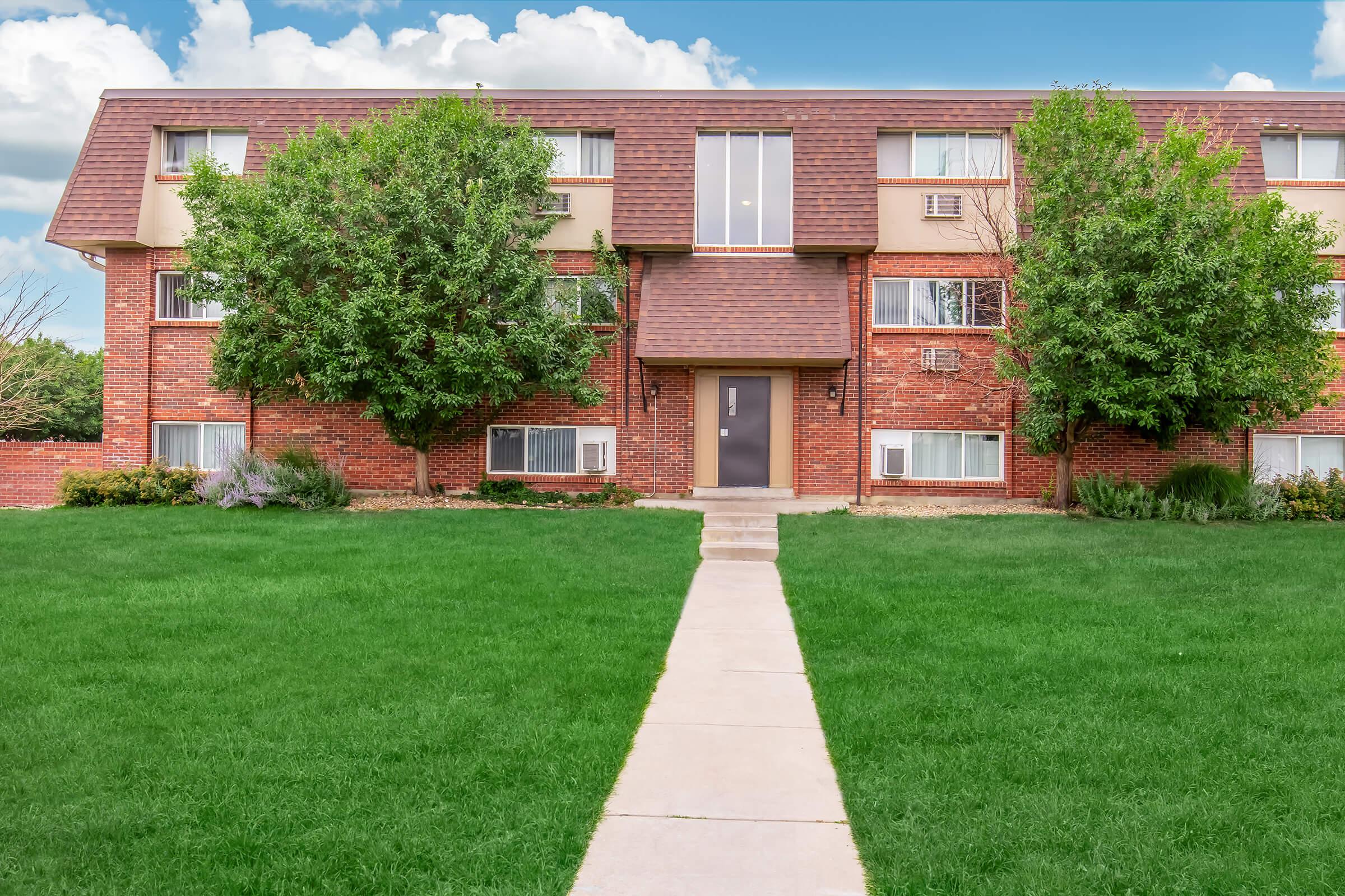 A red brick apartment building with three stories, featuring symmetrical windows and a central entrance. Lush green grass surrounds the building, with a concrete pathway leading to the door. Two trees flank the entrance, and the sky above is partly cloudy.