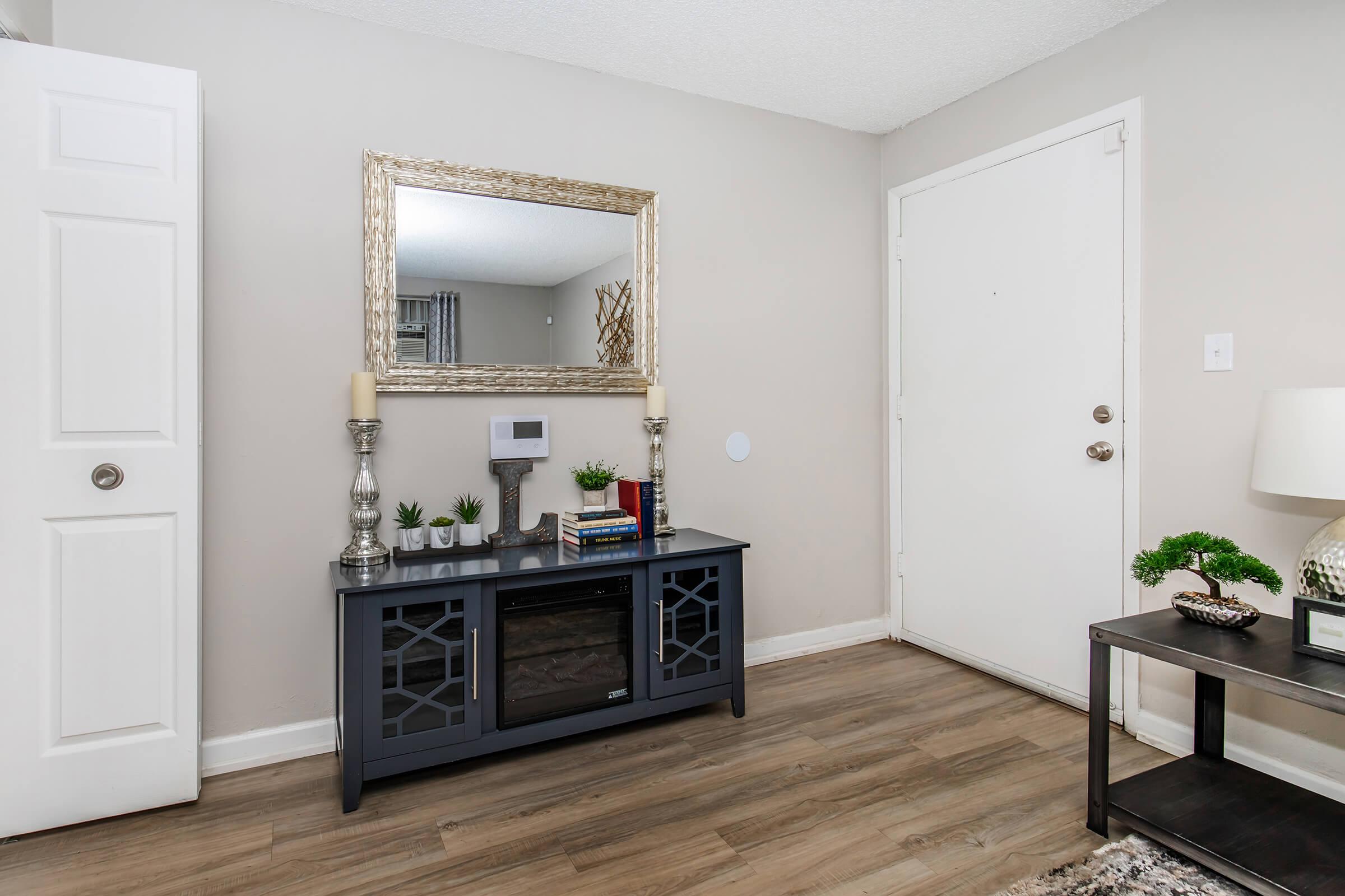 A modern entryway featuring a gray console table with decorative items, including plants and books. Above the table is a large, ornate mirror reflecting the light. To the left, there is a white door. The floor is made of light wood, and a small dark table with a lamp is in the foreground. The walls are painted a soft gray.