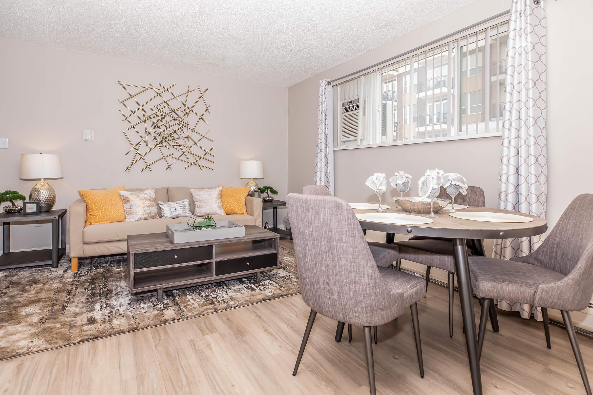 A cozy living and dining area featuring a neutral color palette. The space includes a brown sofa adorned with yellow pillows, a round dining table with gray chairs, and decorative elements like a geometric wall art piece and an air conditioning unit by the window. Soft lighting complements the inviting atmosphere.