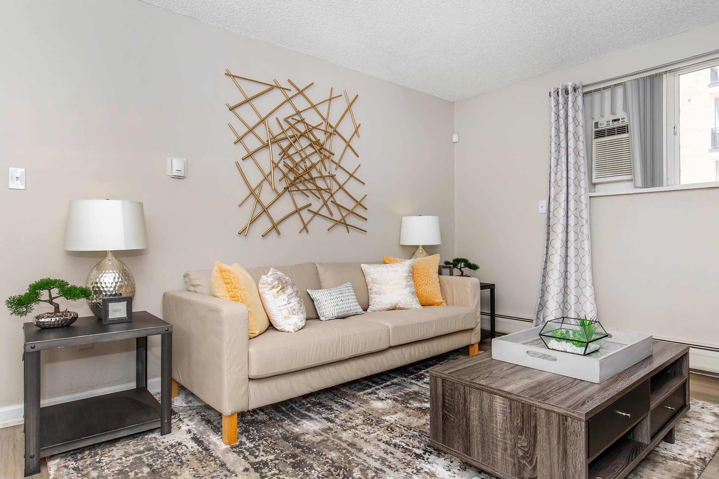 A cozy living room featuring a beige sofa with decorative pillows, two side tables with lamps, and a wooden coffee table with a decorative tray. A wall-mounted piece of art made of bamboo adds interest, while a window allows natural light in, complemented by light-colored walls and a soft area rug.