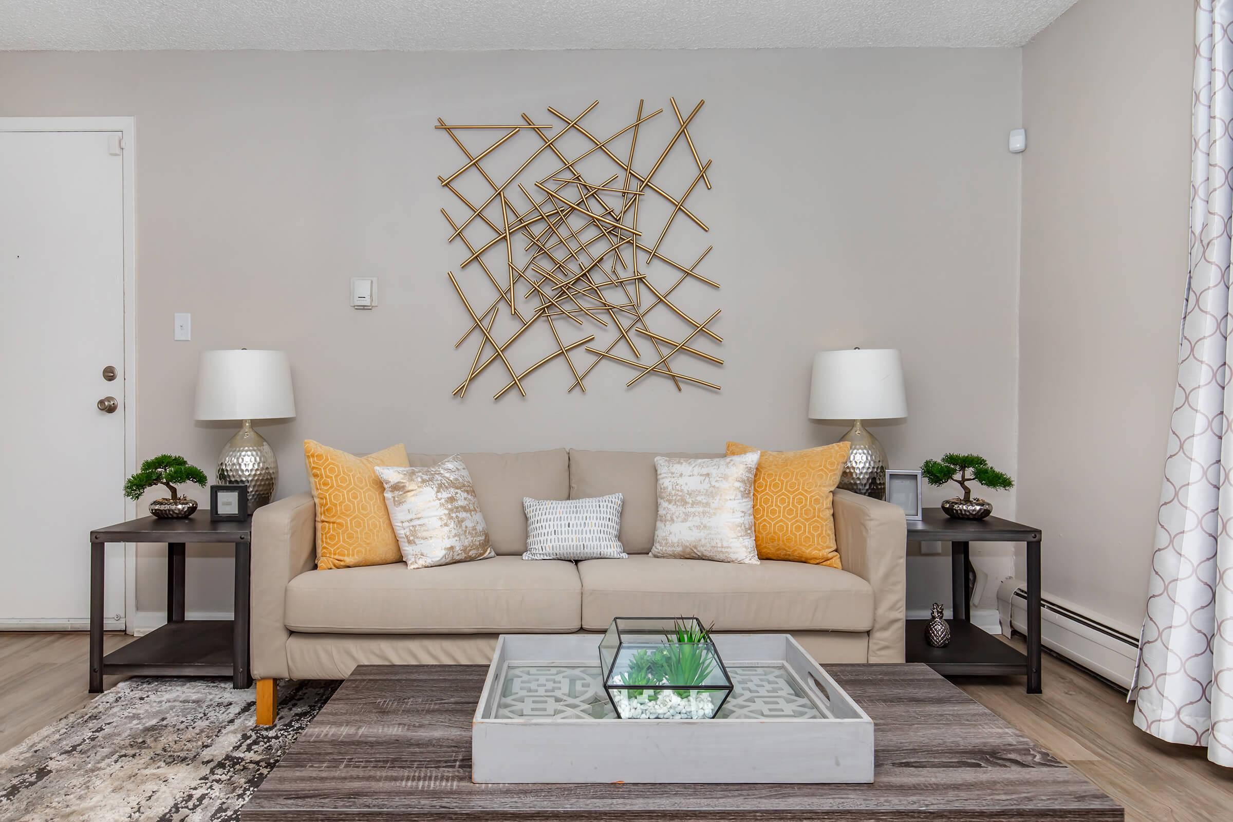 A cozy living room featuring a light beige sofa with decorative pillows in yellow and gray. There are two side tables with lamps and small potted plants on each side. A glass coffee table sits in the center, adorned with a decorative tray and a small plant. The walls are painted a neutral color, and a modern art piece hangs above the sofa.