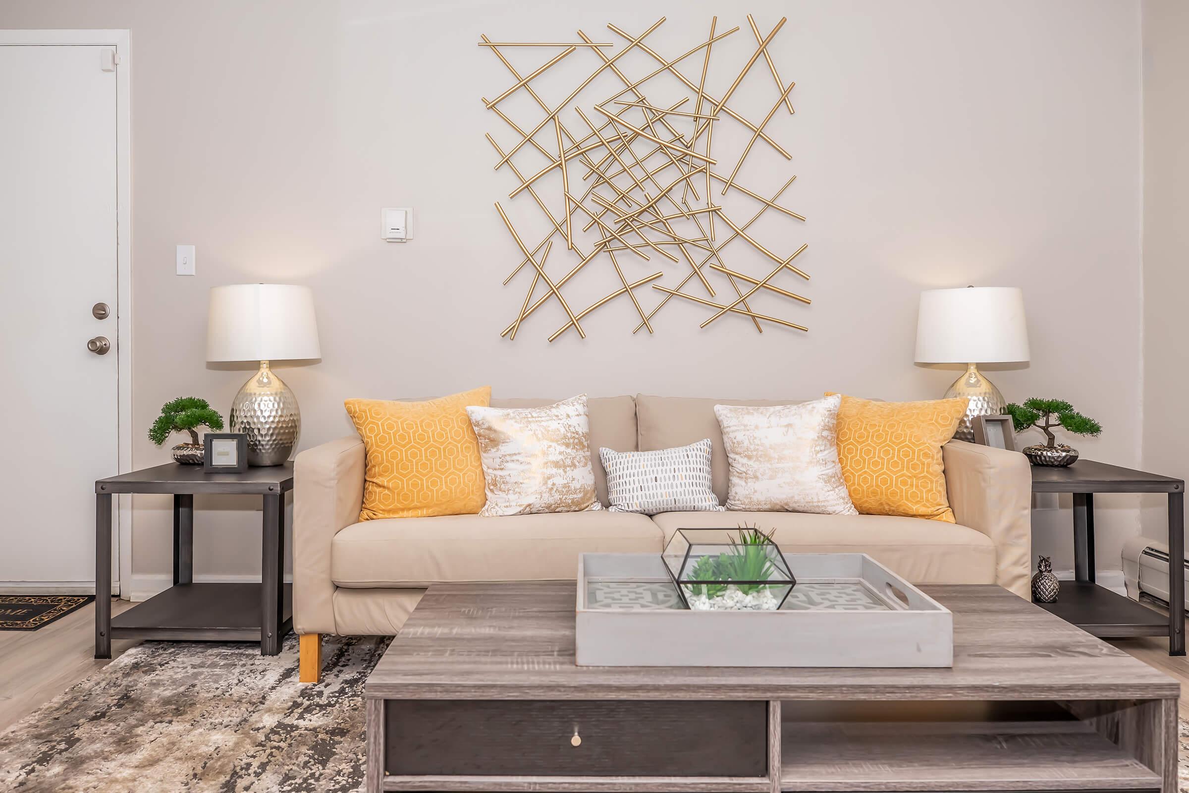 A cozy living room featuring a light-colored sofa adorned with decorative yellow and patterned pillows. Two stylish side tables with lamps flank the sofa. A decorative metal wall art piece is displayed above the sofa, and a coffee table with a glass centerpiece sits in front, enhancing the warm and inviting atmosphere.