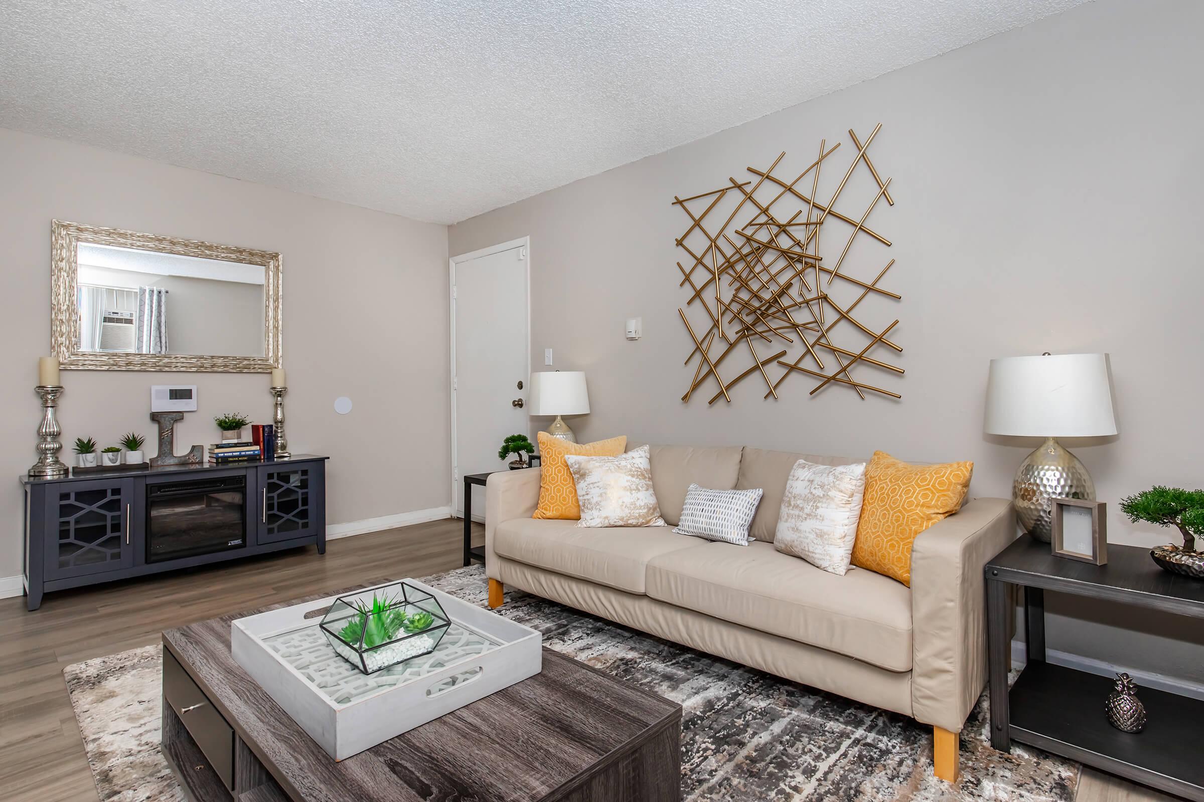 A stylish living room featuring a beige sofa with decorative pillows, a coffee table with a plant centerpiece, a dark wood media console, and wall decor. There are two lamps on side tables, a large mirror, and textured wall accents, creating a cozy and modern atmosphere.