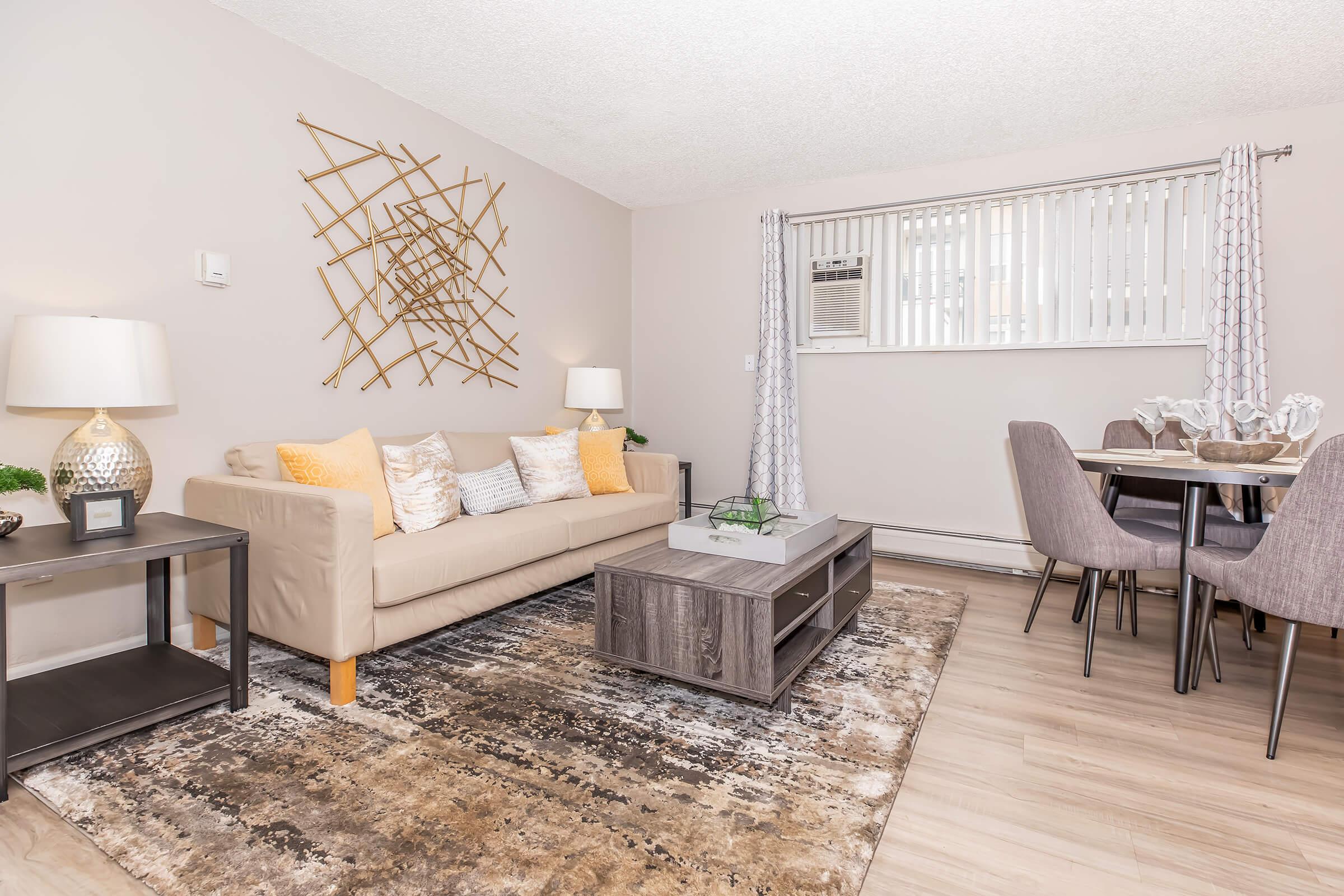 A cozy living room featuring a light-colored sofa with decorative pillows, a coffee table, and a stylish area rug. To the side, a dining table with gray chairs is visible, along with a wall-mounted air conditioning unit and elegant wall decor. Natural light streams in through the window.