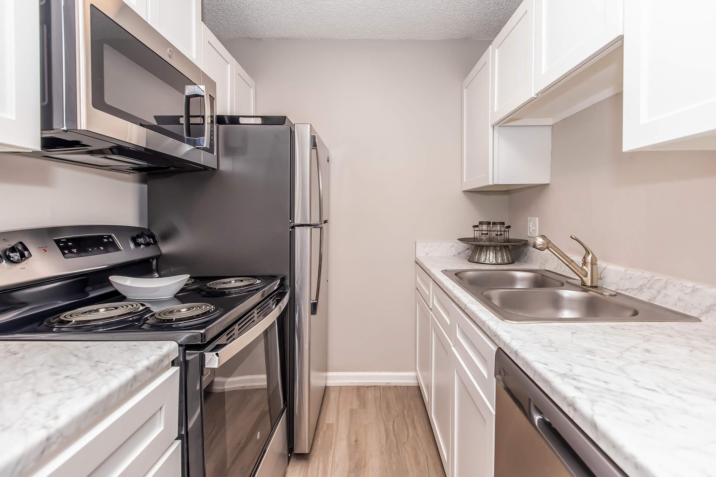 A modern kitchen featuring stainless steel appliances including a microwave, refrigerator, and stove. The countertops are made of marble, and there are white cabinetry and a double sink. The flooring is light-colored, enhancing the contemporary look of the space.