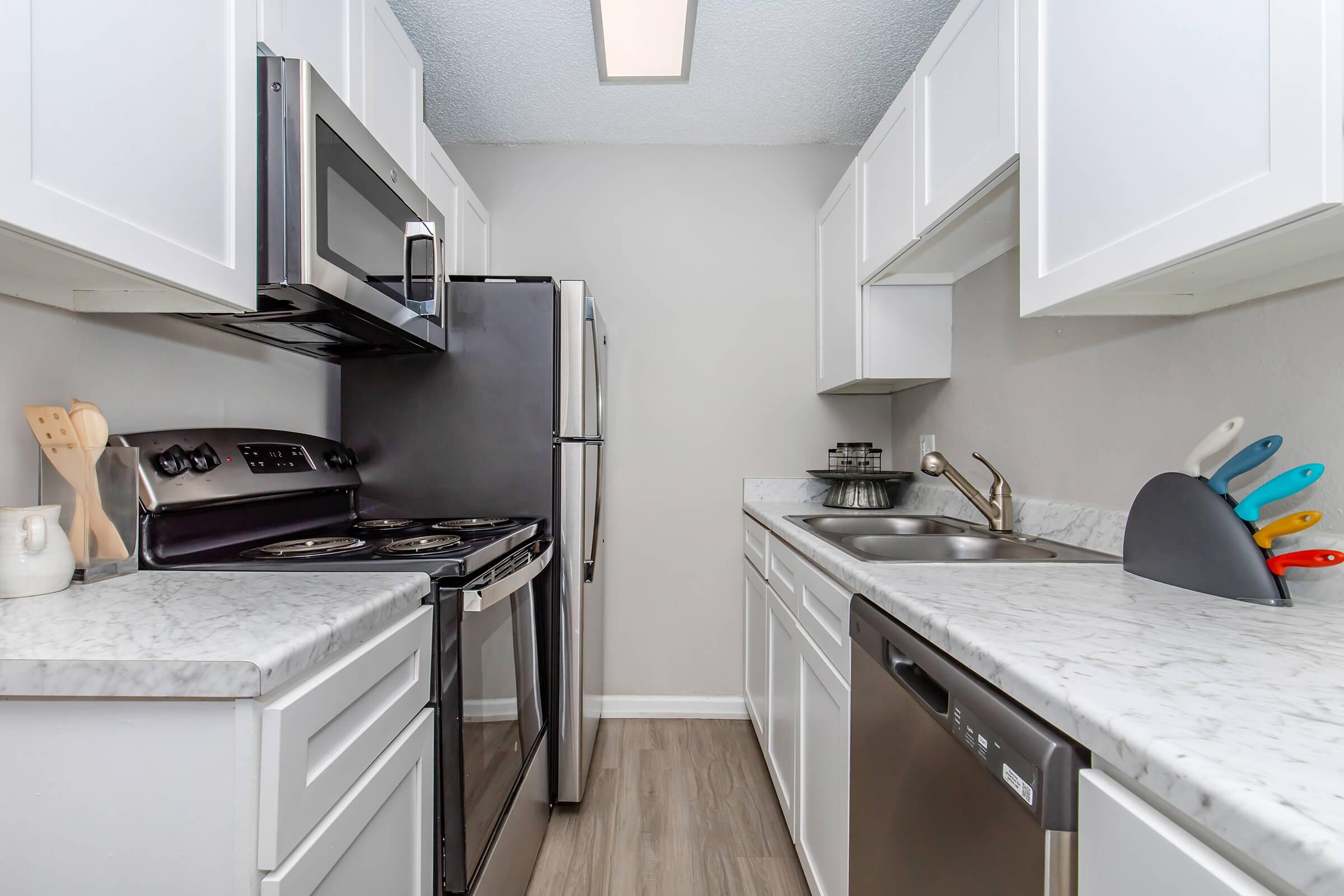 Modern kitchen featuring white cabinets, a black refrigerator, stainless steel appliances, and a marble countertop. The kitchen includes a dual sink, a stove with an oven, and a dishwasher, with colorful kitchen utensils displayed in a holder. The overall design is clean and contemporary.