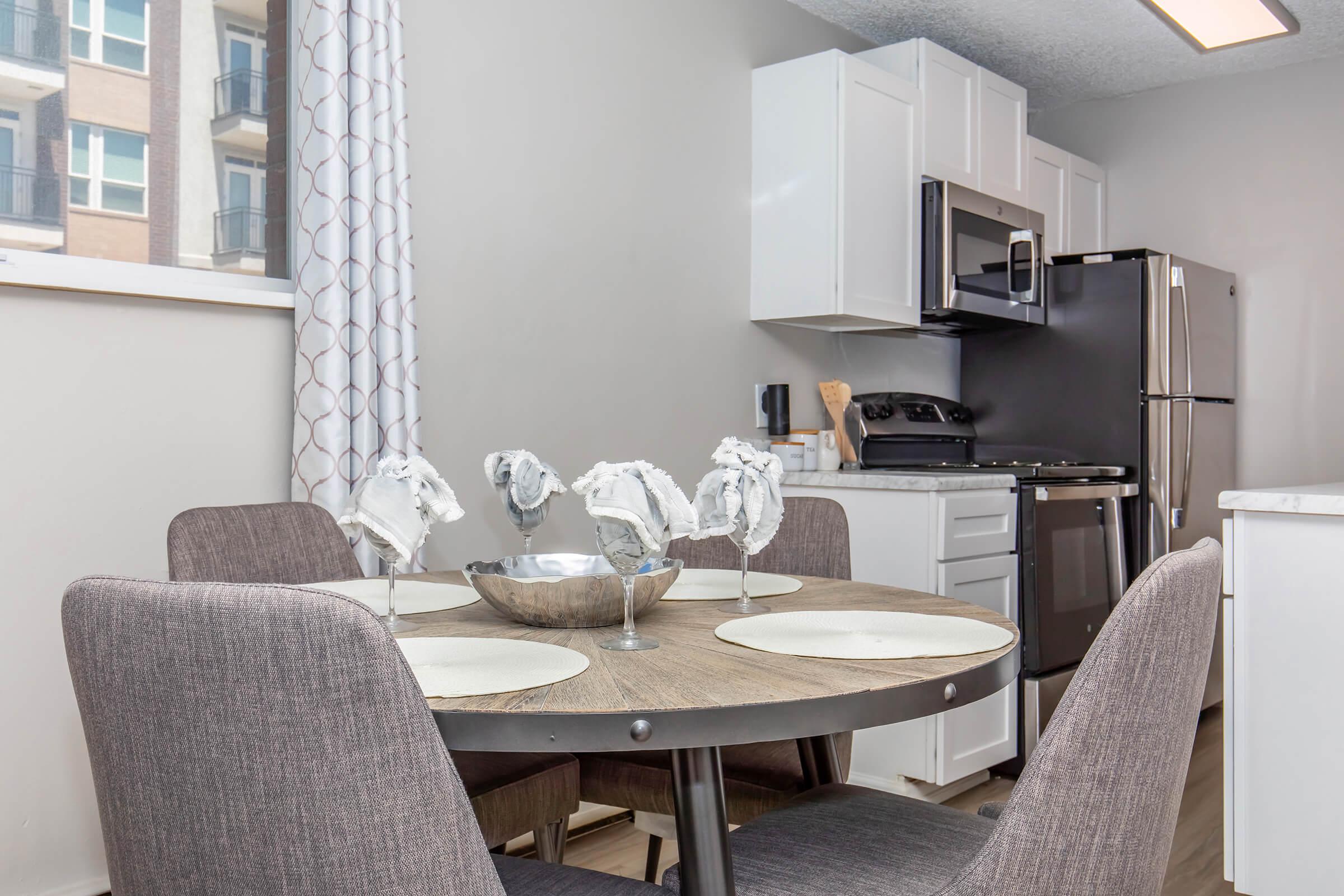 A modern kitchen featuring a round wooden table set with four gray chairs, a decorative bowl in the center, and place settings. The kitchen includes white cabinets, stainless steel appliances, and a large window with light gray curtains. The space is bright and inviting, showcasing contemporary design elements.