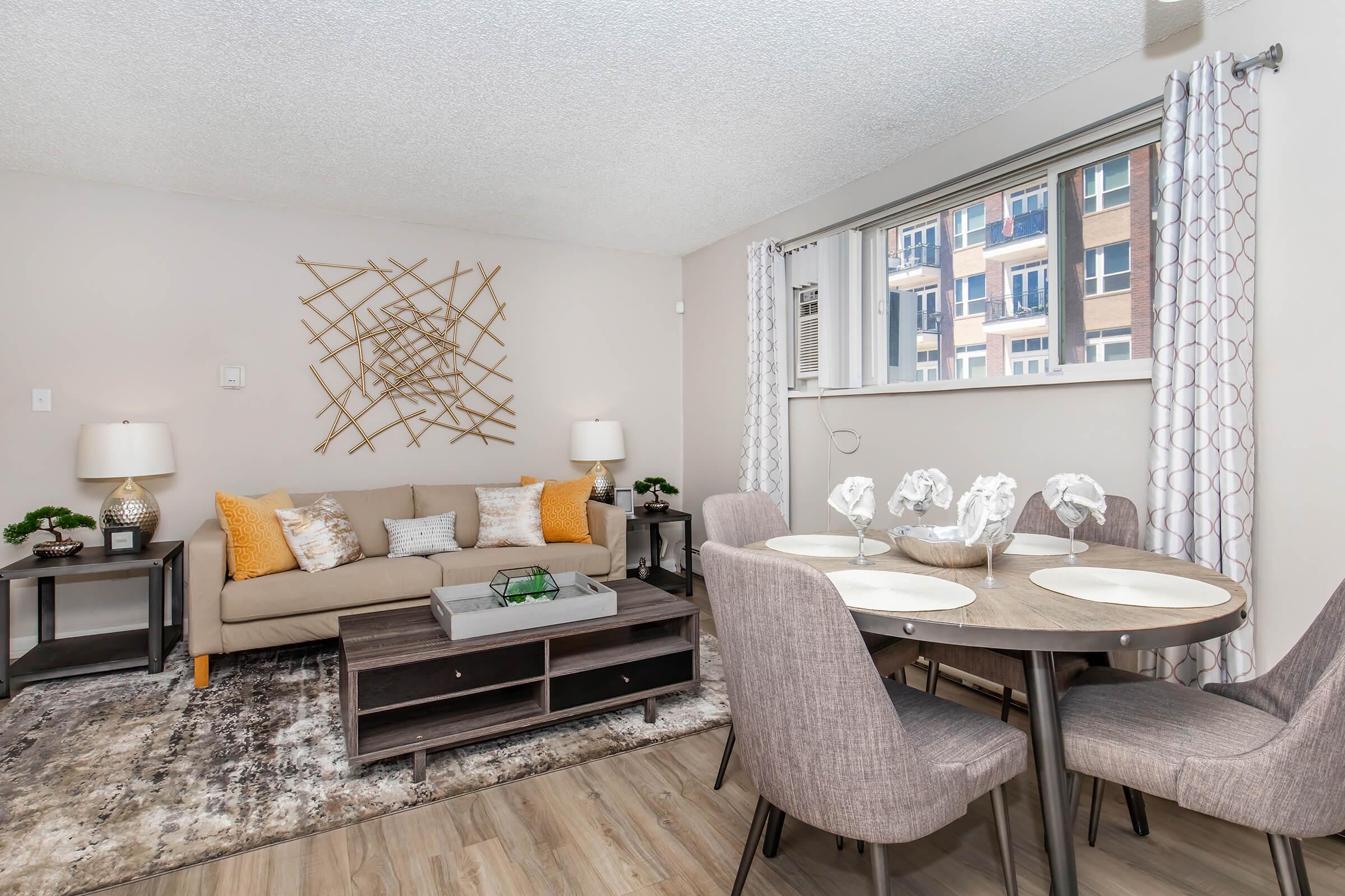 A modern living room and dining area featuring a light-colored sofa with decorative pillows, a round dining table set for four, and stylish lamps on side tables. The walls are adorned with a geometric wall art piece, and natural light filters in through large windows, offering a cozy and inviting atmosphere.