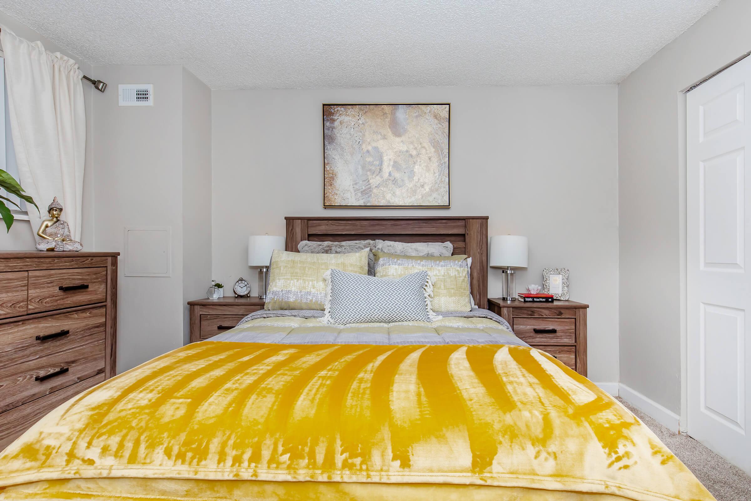 A cozy bedroom featuring a queen-sized bed with a yellow and white striped throw blanket. The bed is flanked by two nightstands with lamps. A wooden dresser is on the left, and a piece of abstract art hangs above the bed. The walls are painted a soft neutral color, creating a calming atmosphere.