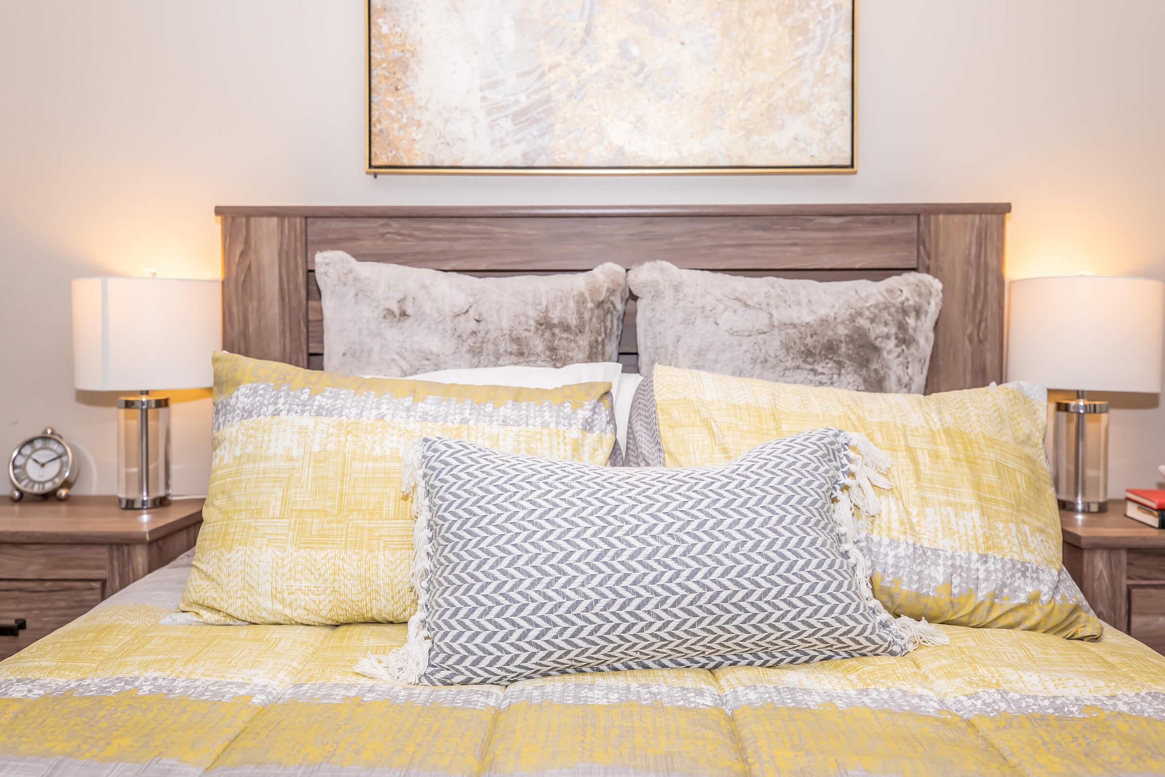 A cozy bedroom scene featuring a wooden headboard with decorative pillows in yellow and gray hues on a neatly made bed. The room is softly illuminated by two bedside lamps, and a piece of abstract art hangs above the bed, adding a warm, inviting atmosphere.