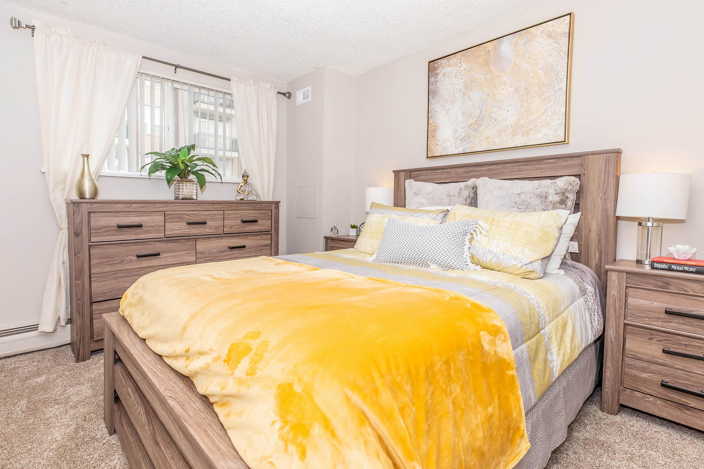 A cozy bedroom featuring a wooden bed frame with a yellow and gray bedspread, matching pillows, and two bedside lamps. A wooden dresser is positioned against the wall, and a large piece of abstract art hangs above the bed. Natural light filters in through a window adorned with white curtains.