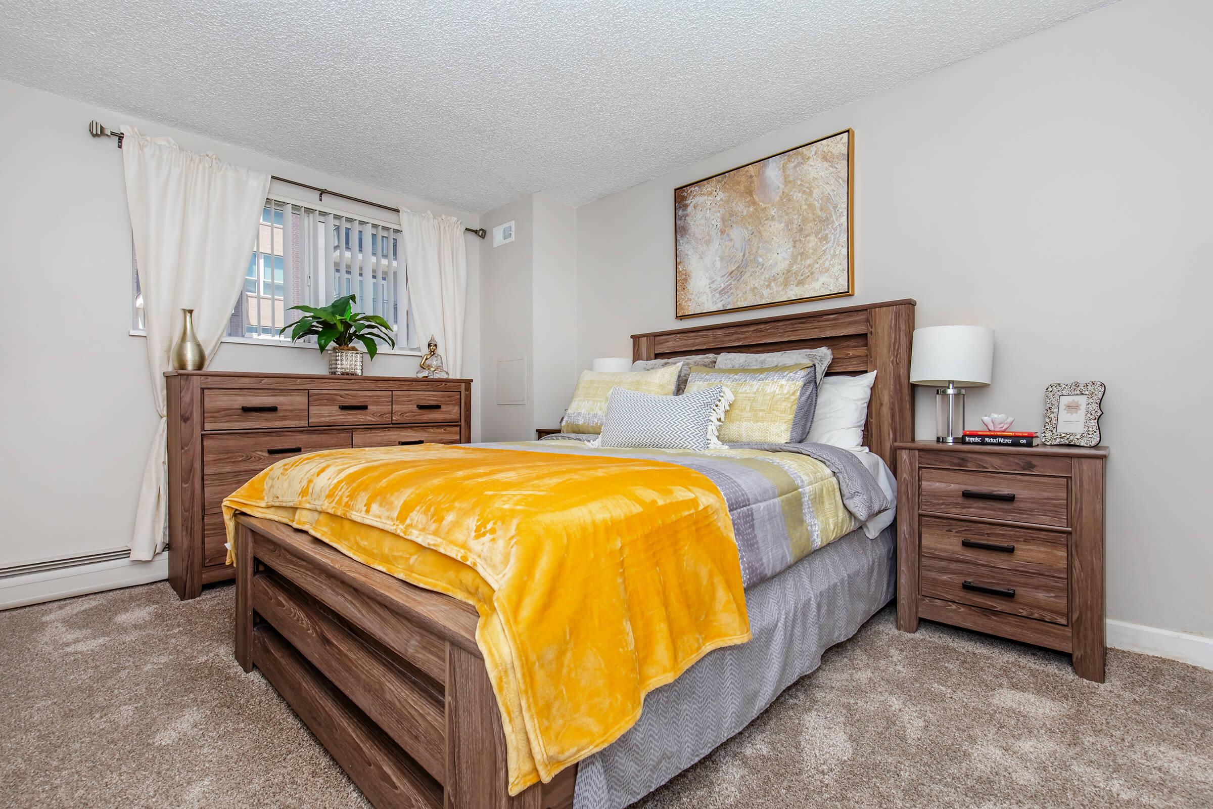 A cozy bedroom featuring a wooden bed frame with a yellow and gray blanket, paired with matching nightstands and a dresser. The room has light-colored walls, soft carpeting, and large windows with sheer curtains letting in natural light. A piece of abstract art hangs above the bed, and a potted plant adds a touch of greenery.