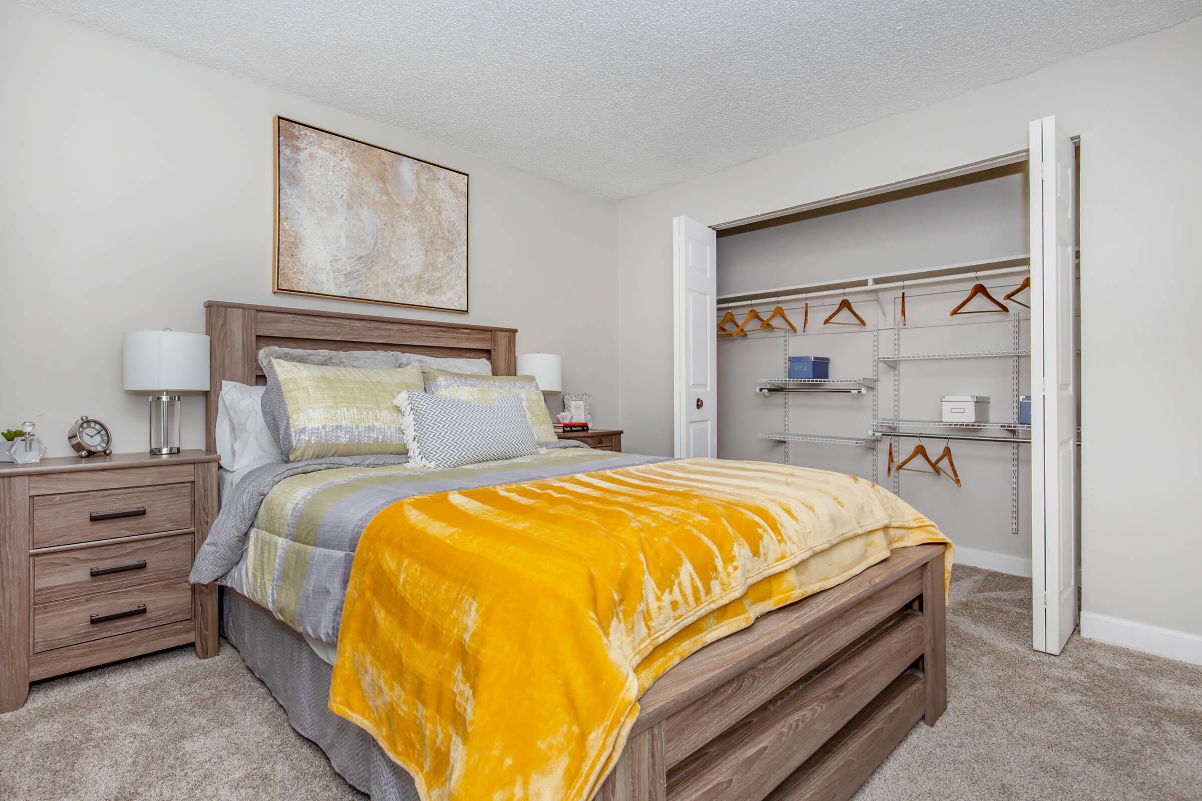 A cozy bedroom featuring a wooden bedframe with a yellow and gray blanket, bedside tables with lamps, and a piece of abstract wall art. There is an open closet with hanging space and shelves, revealing additional storage options. The room has neutral-colored walls and carpet.