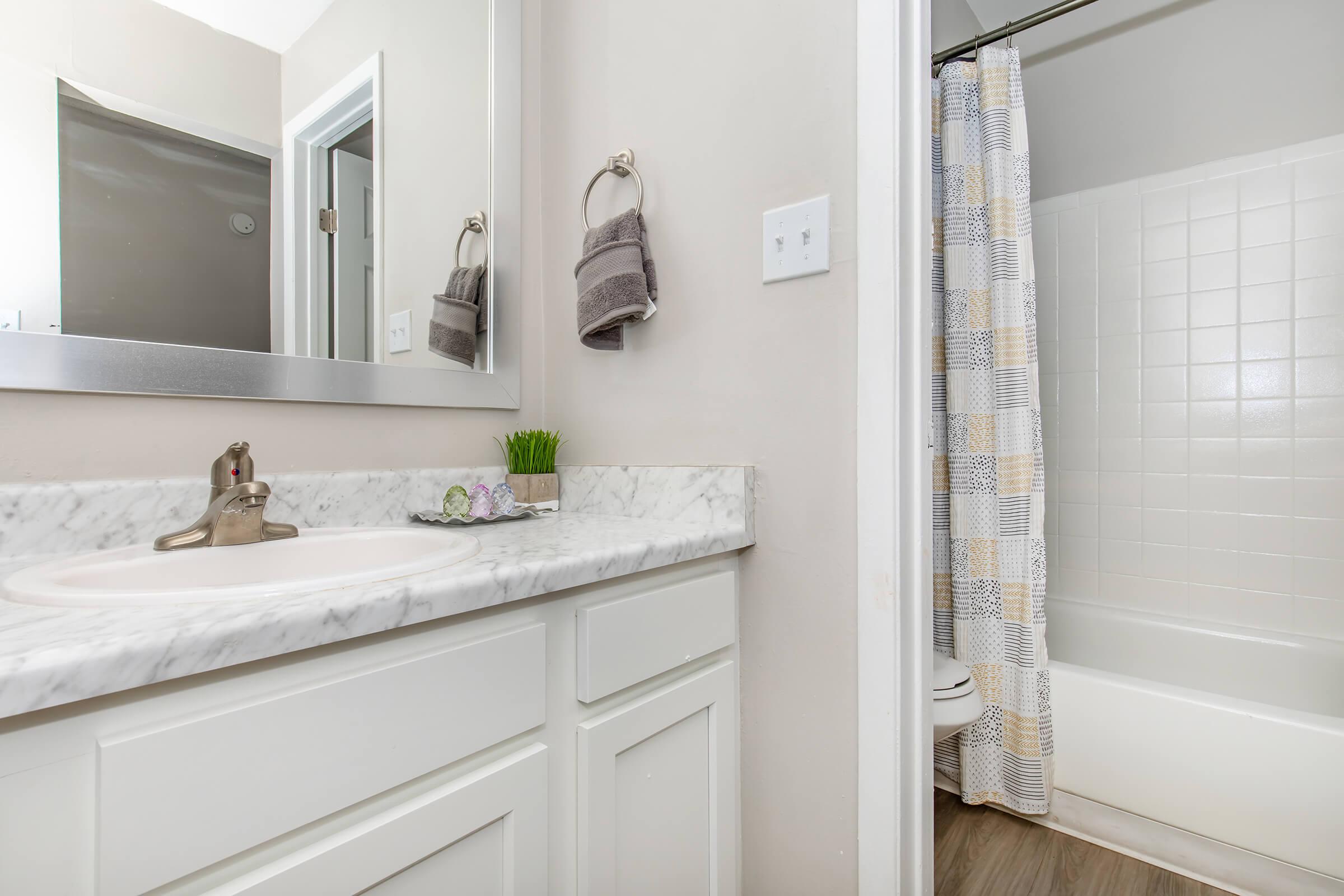 A modern bathroom featuring a white marble countertop with a sink, a mirror on the wall, a decorative plant, and gray towels hanging on towel bars. In the background, there's a bathtub with a shower curtain in a geometric pattern, creating a clean and inviting atmosphere.