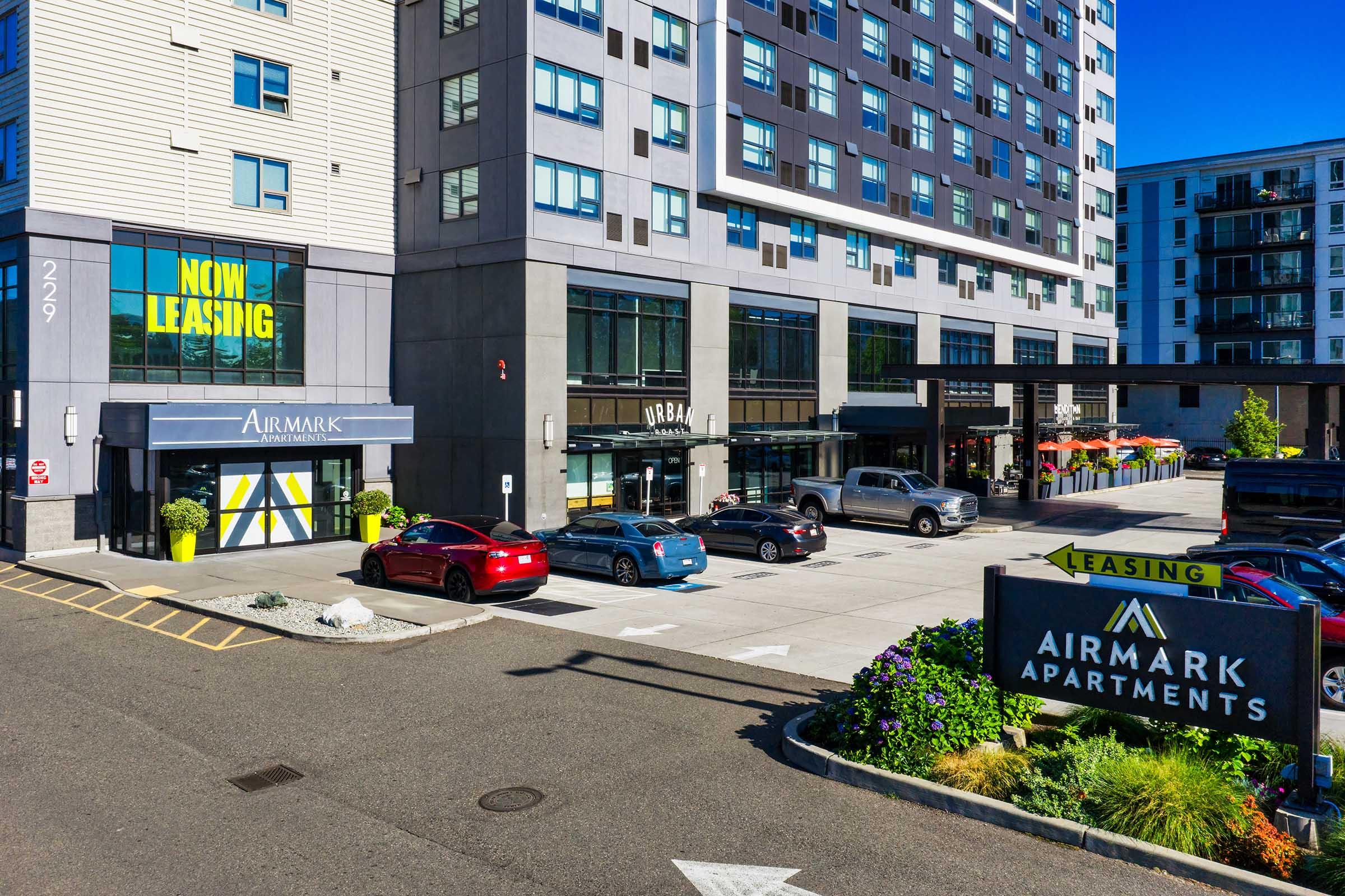Exterior view of Airmark Apartments building featuring a leasing office with a marquee sign reading "NOW LEASING," surrounded by landscaped greenery. Parking area with several cars is visible. The complex has modern architecture and a welcoming entrance for potential residents.