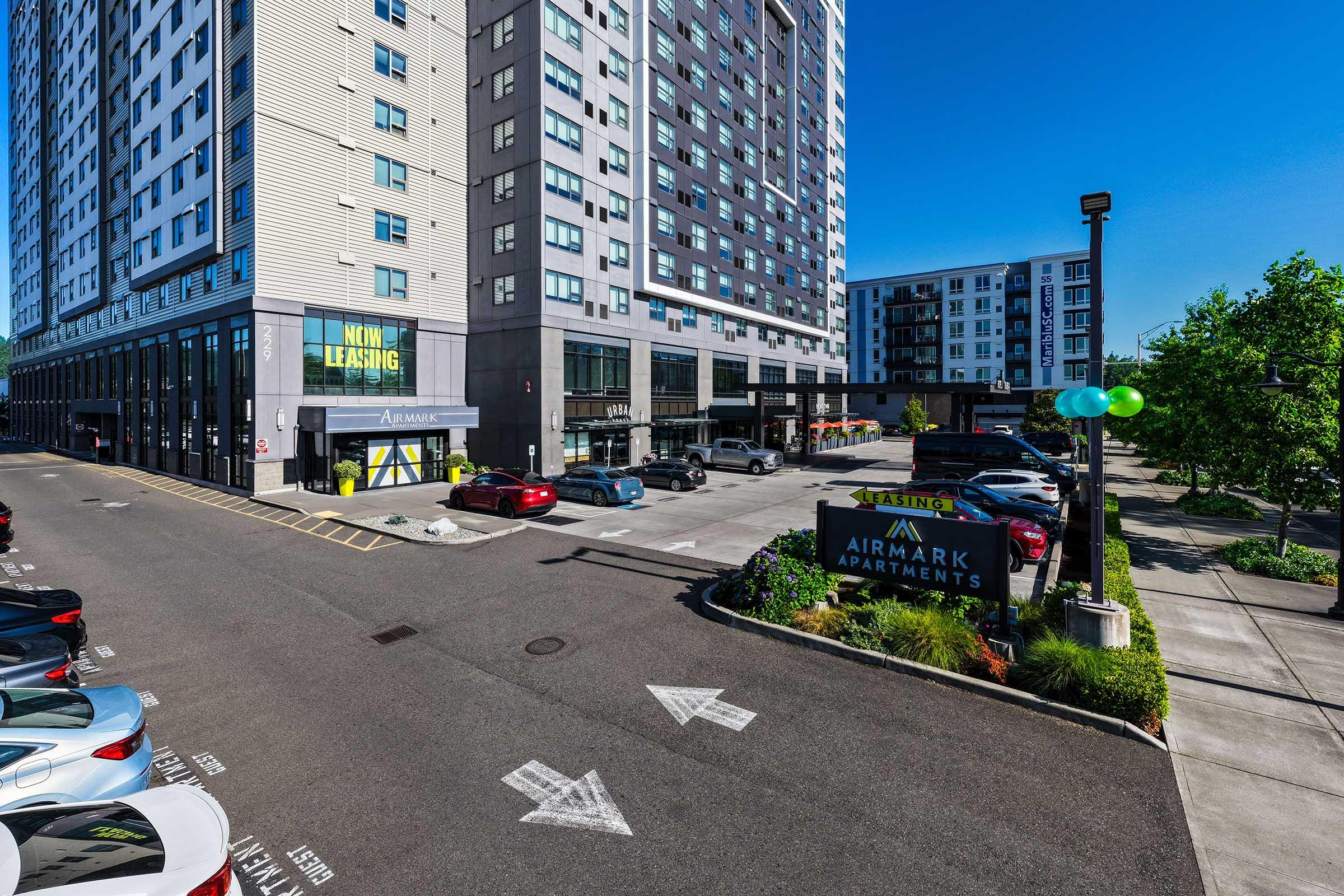 Exterior view of a modern apartment complex called Airmark Apartments. The building features large windows and a leasing office with a prominent "Now Leasing" sign. The parking lot is lined with cars, and there are green balloons adorning a sign. The sky is clear and blue, contributing to the bright, inviting atmosphere.