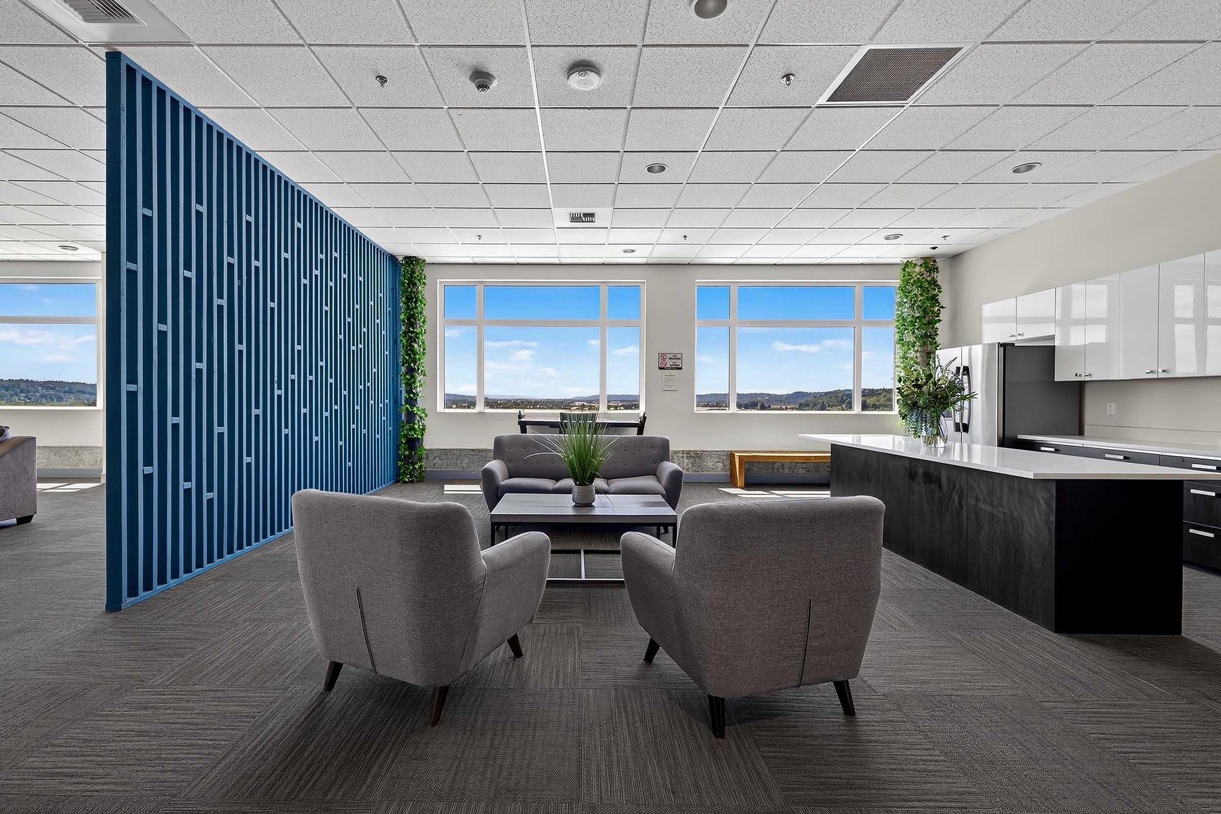 A modern office lounge with two gray armchairs facing a small coffee table. Behind is a blue decorative wall, and large windows showcase a scenic view. The room features a kitchenette with sleek cabinetry and plants for added aesthetics. The floor is dark carpeted, creating a cozy atmosphere.