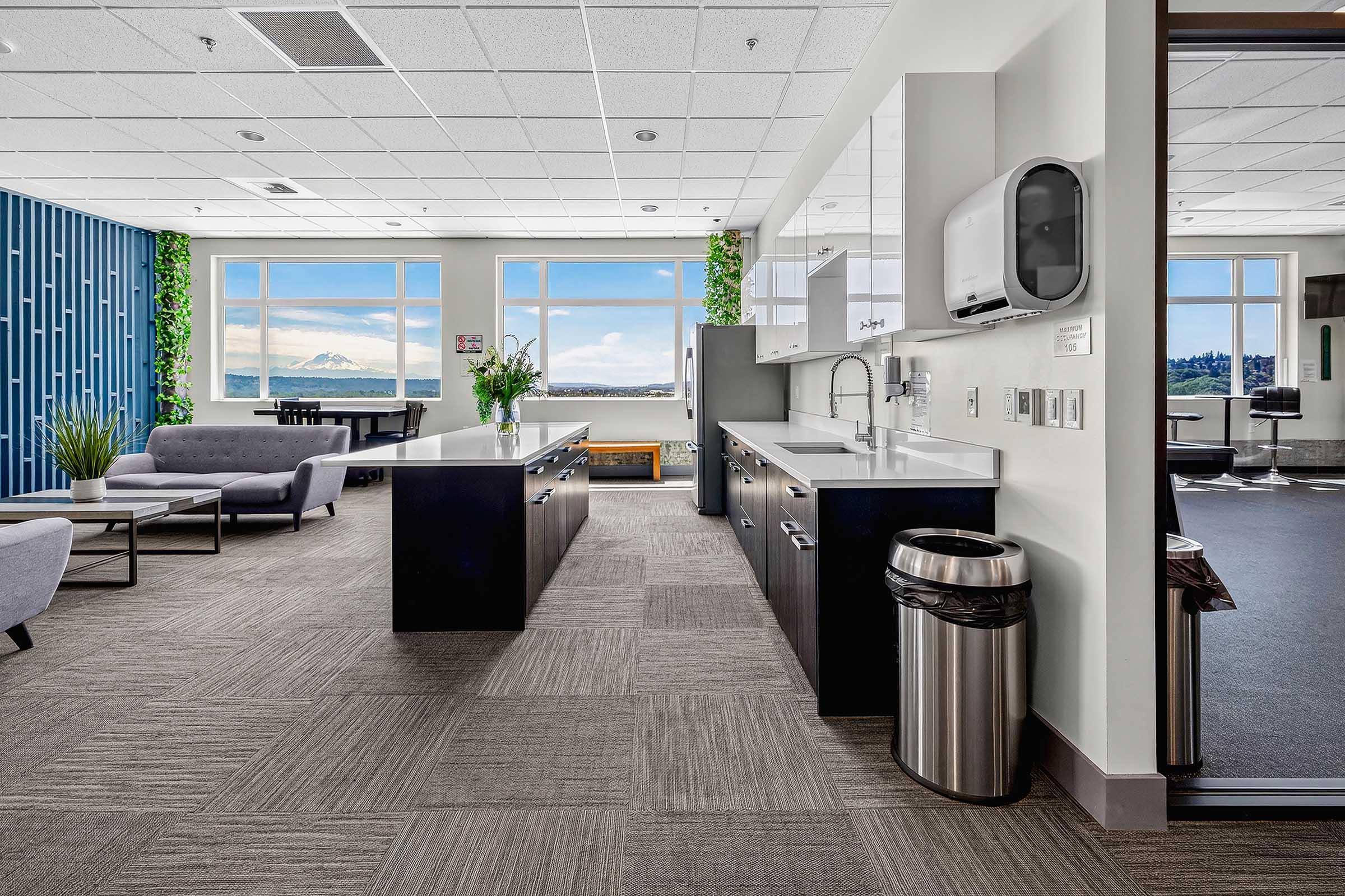 Modern office kitchen area featuring a large island with cabinets and sink, lounge seating, and large windows with a view. Potted plants add greenery, and there's a trash can visible. The space is well-lit and contemporary, designed for relaxation and work.