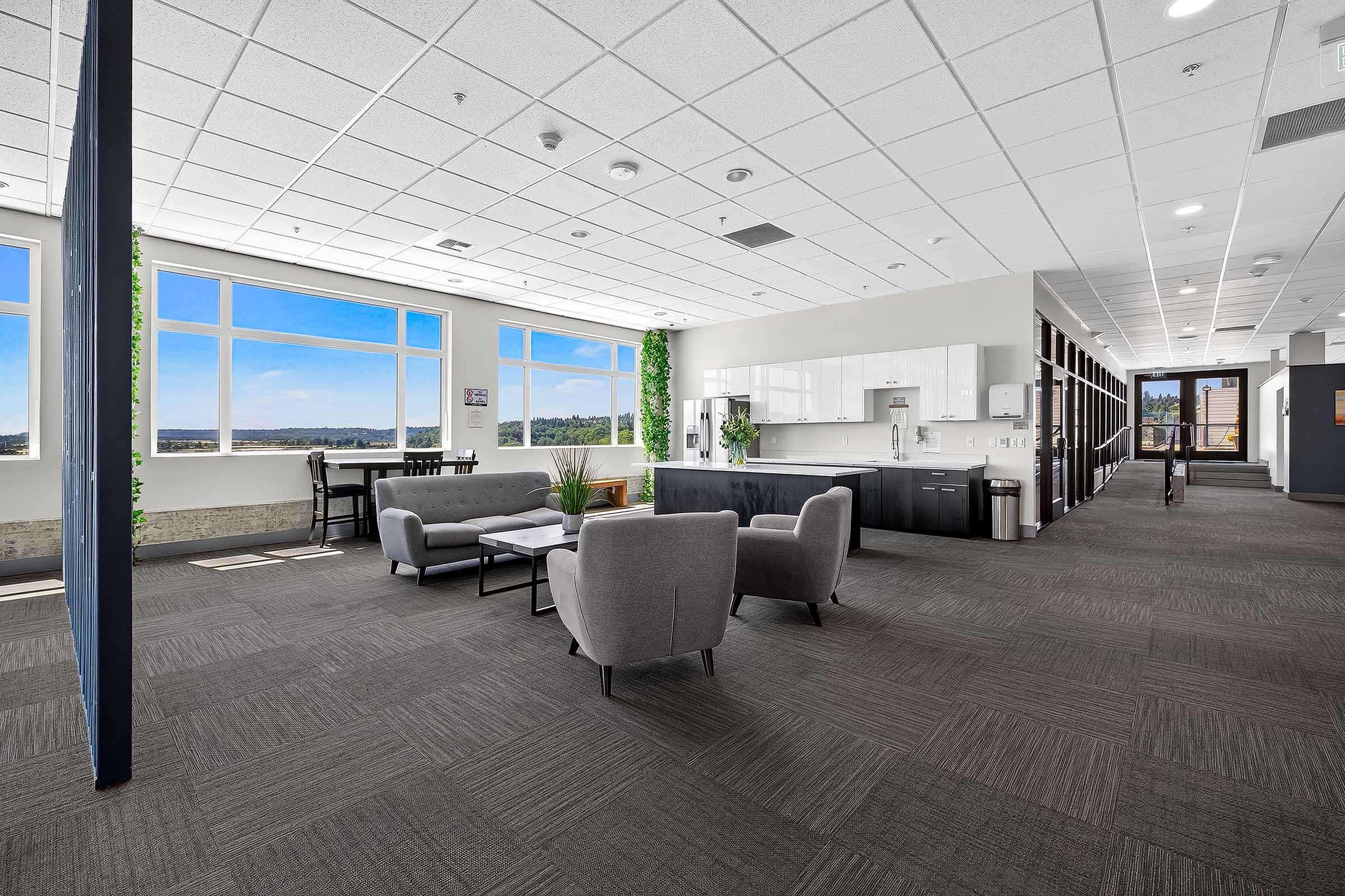 A modern office interior featuring a spacious break area with large windows, comfortable gray seating arrangements, a small coffee table, and a kitchenette. The space has neutral-colored walls and carpeting, complemented by plants and ample natural light, creating a bright and inviting atmosphere.