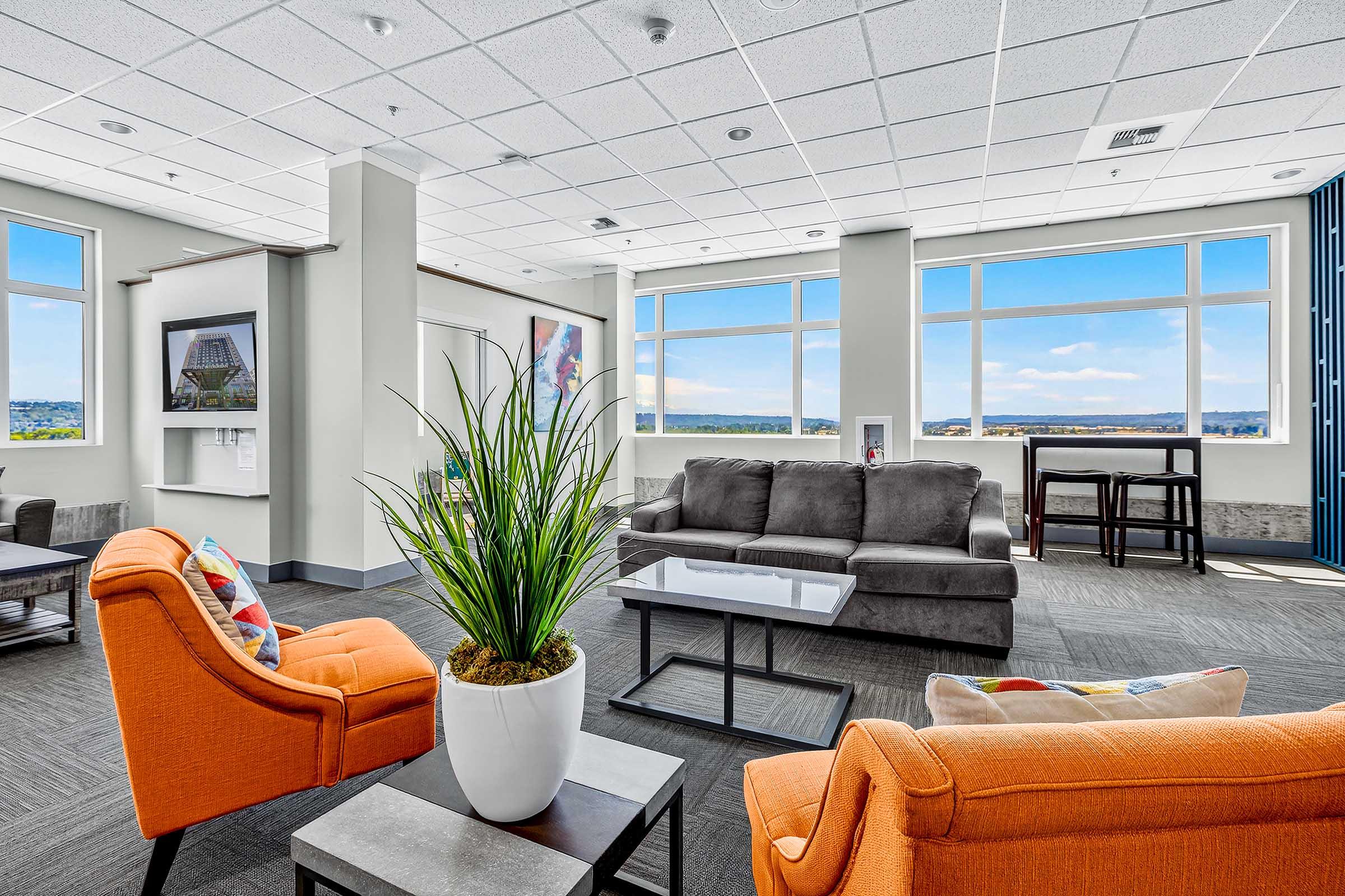 A modern, bright lounge area featuring orange armchairs, a gray sofa, and a coffee table. Large windows provide a panoramic view of the outdoors, with blue skies and distant scenery. Decorative plants add warmth to the contemporary design, creating an inviting atmosphere for relaxation or socializing.