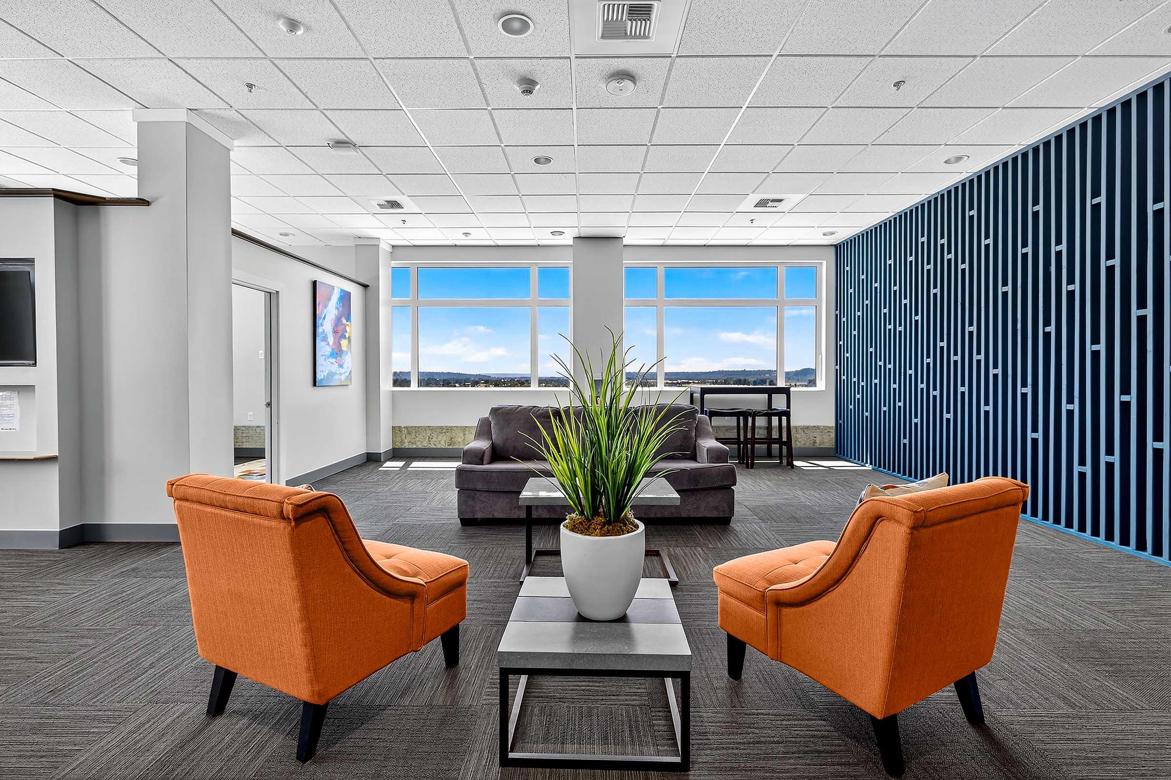 A modern lounge area featuring two orange chairs and a gray couch. There's a small table between the chairs with a potted plant. Large windows offer a view of the outdoors, and a blue accent wall adds a stylish touch to the space. A television is mounted on one wall, enhancing the inviting atmosphere.