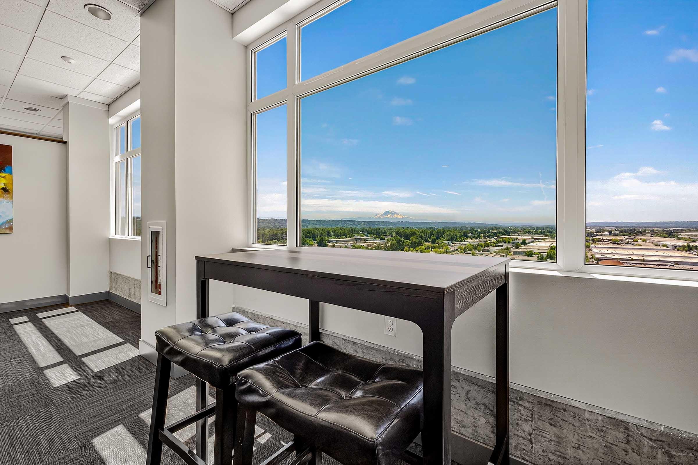 A modern interior space featuring a high table with two black stools, positioned near large windows. The view beyond showcases a clear sky and mountainous landscape, with industrial and urban scenery below. Bright natural light illuminates the room, highlighting its contemporary design.