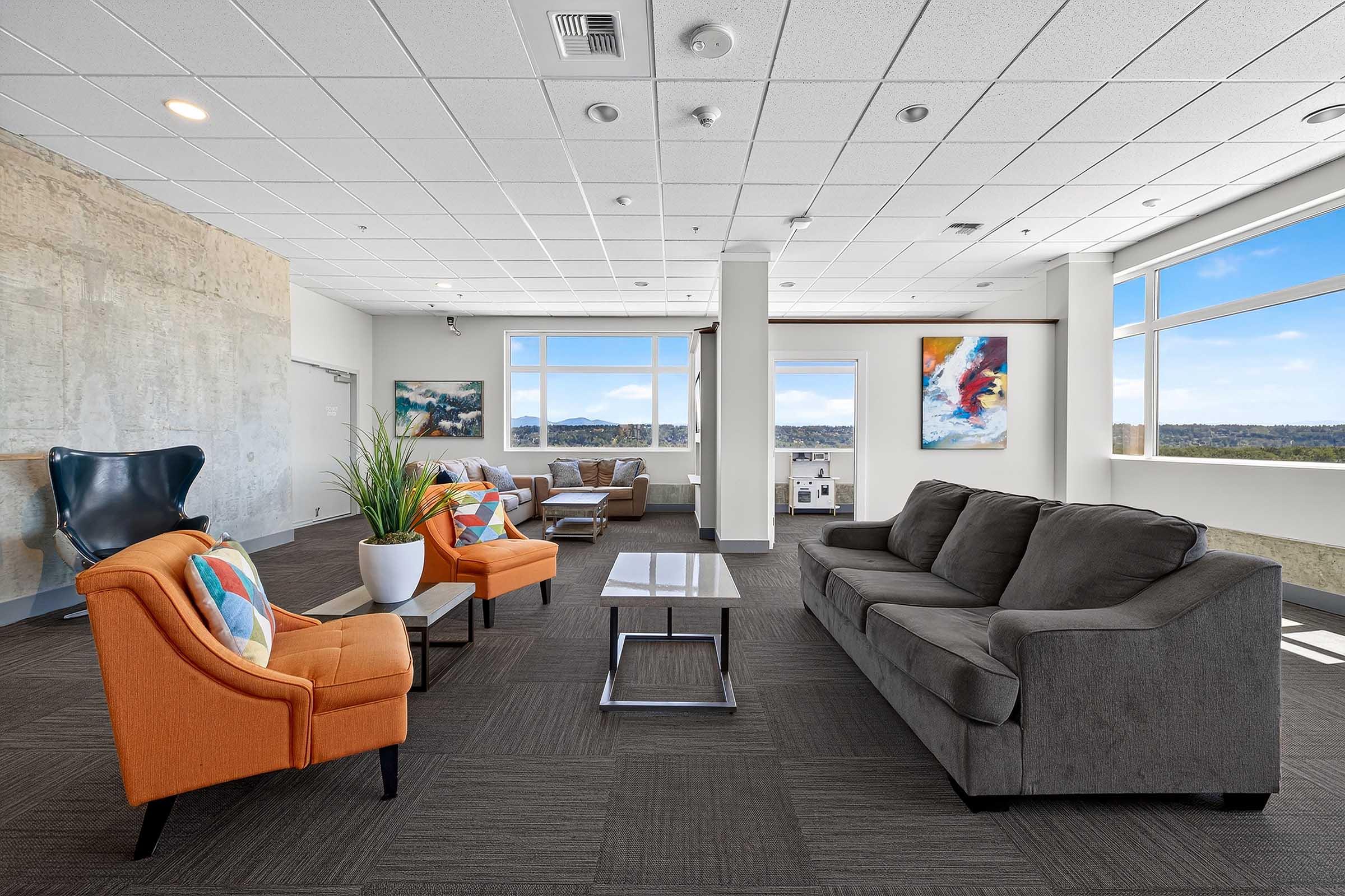 Modern lounge area featuring a gray sofa, orange accent chairs, and a glass coffee table. Large windows provide natural light and a scenic view. Decor includes abstract artwork on the walls and potted plants, creating a comfortable and inviting atmosphere.