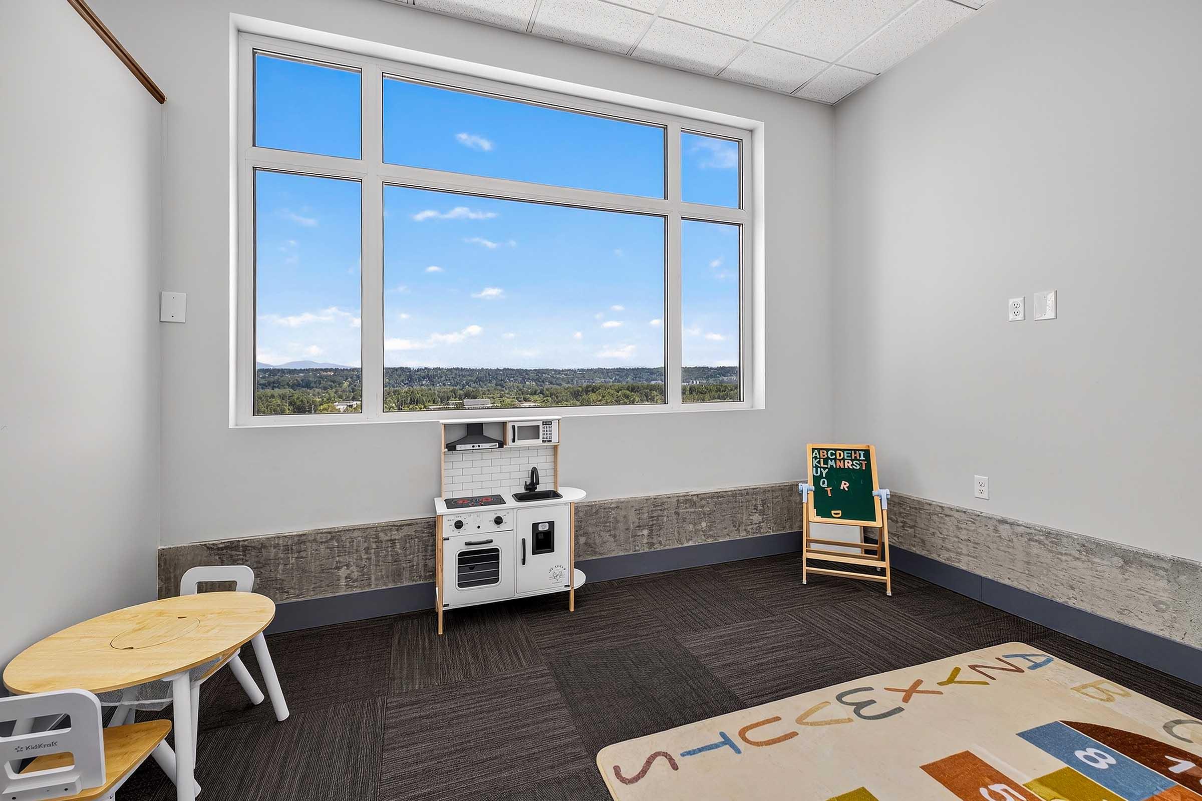 A bright, minimalistic playroom featuring a small wooden table and chairs, a play kitchen set against a wall, a colorful rug, and a chalkboard. Large windows allow natural light and offer a view of green hills and blue sky. The walls are painted in neutral tones, creating a welcoming atmosphere.