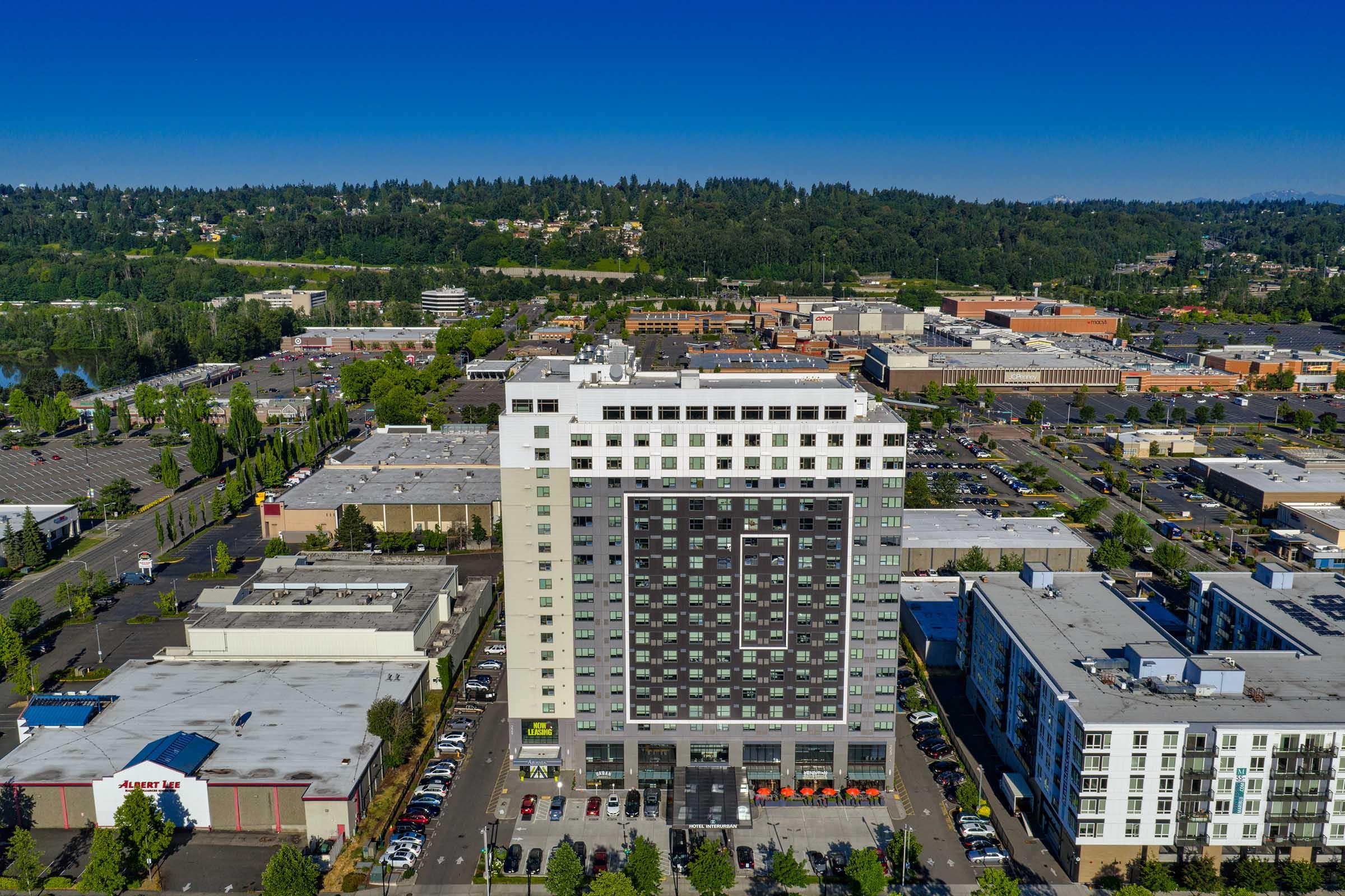 Aerial view of a modern high-rise building surrounded by commercial buildings and parking lots. The building features a mix of dark and light exterior designs, with multiple windows. Lush green hills are visible in the background, indicating a suburban environment. Clear blue sky enhances the overall scene.