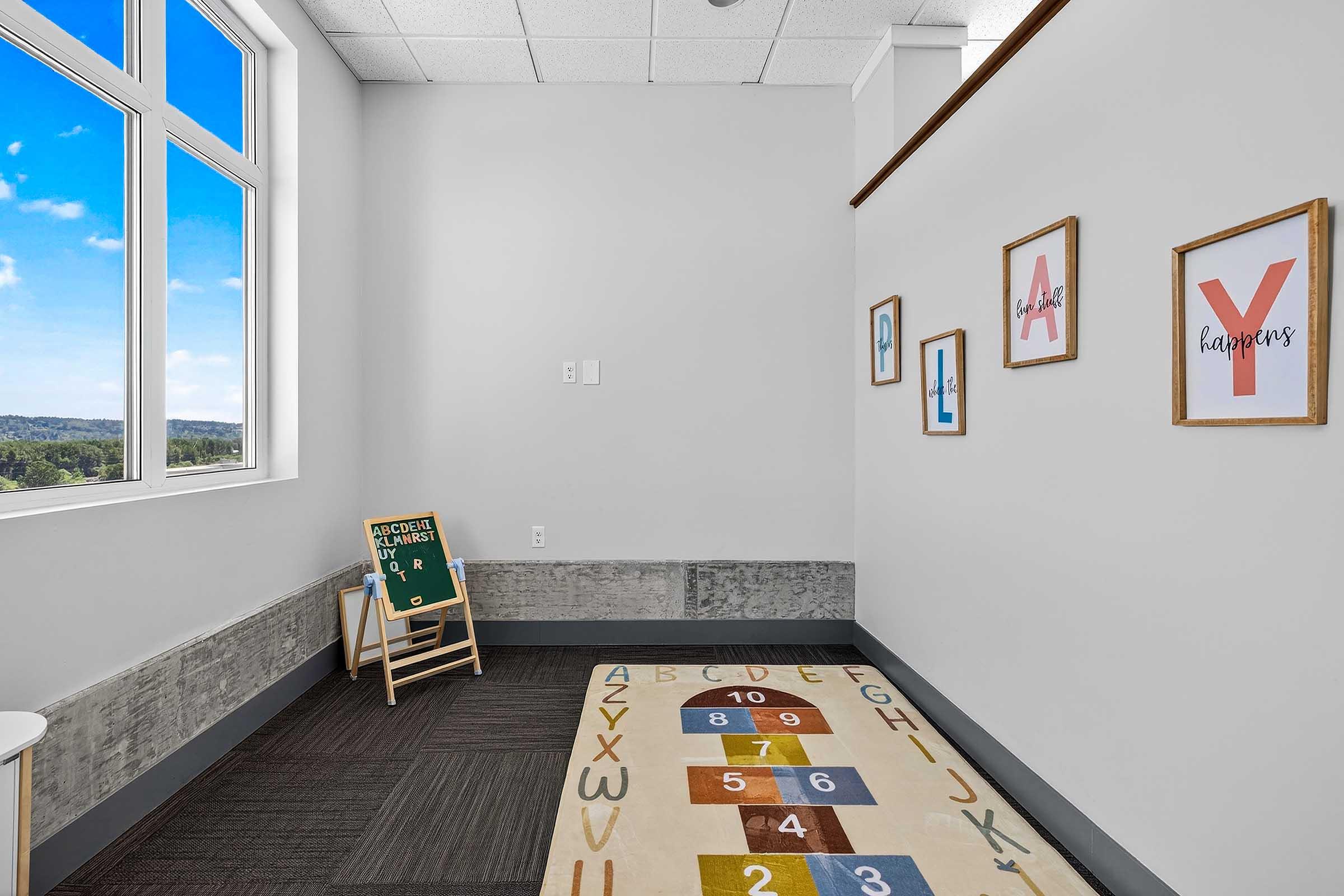 A bright corner of a room featuring a hopscotch rug on the floor, a small chalkboard with a welcoming message, and framed letters on the wall. Large windows offer a view of the outdoors, flooding the space with natural light.