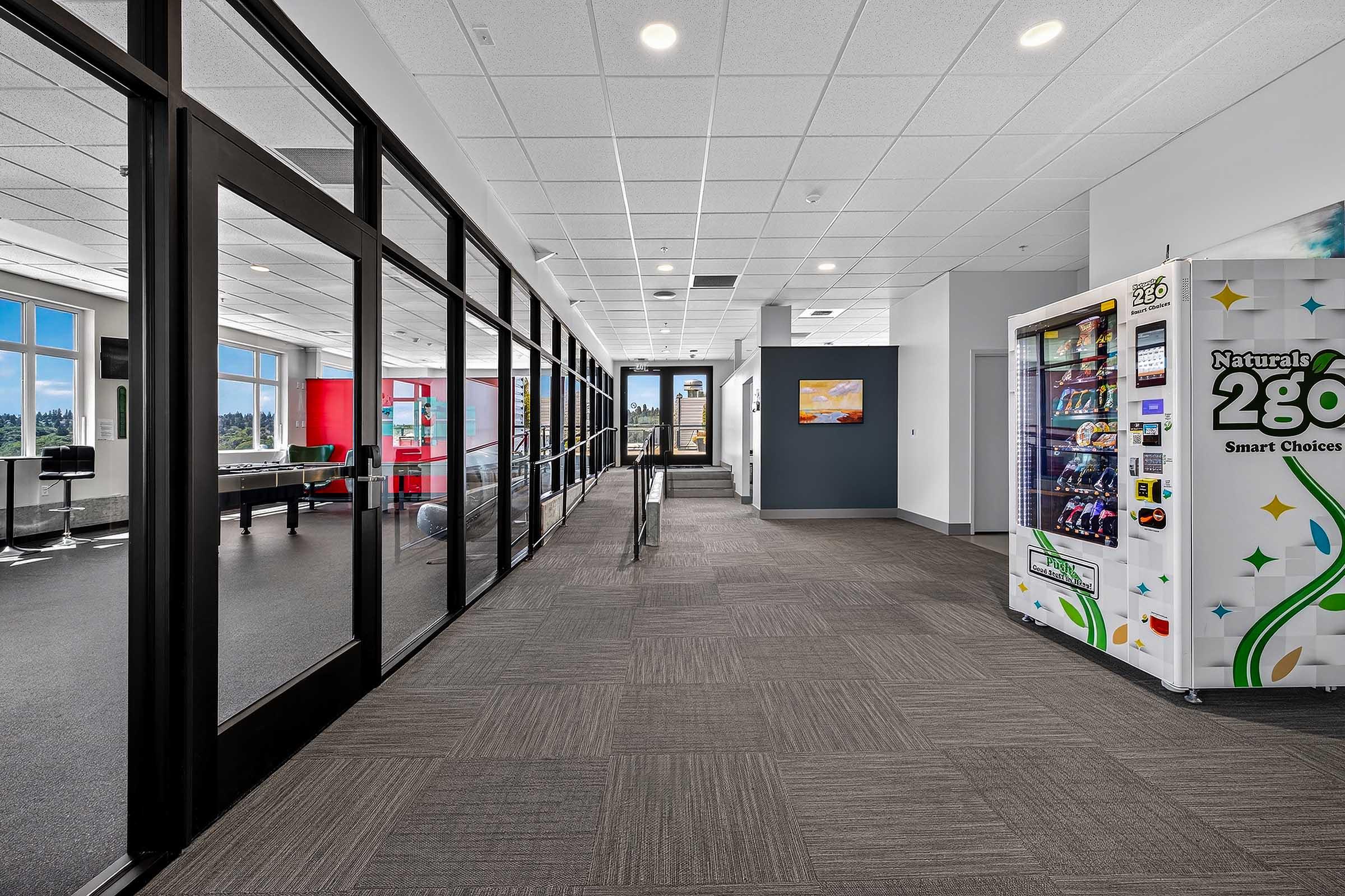 A modern office hallway with large windows allowing natural light. On one side, there are glass walls leading to meeting rooms, while on the other, a vending machine labeled "Natural's 280 Smart Choices." The floor is carpeted in a gray pattern, and there are colorful accents on the walls, enhancing the inviting atmosphere.