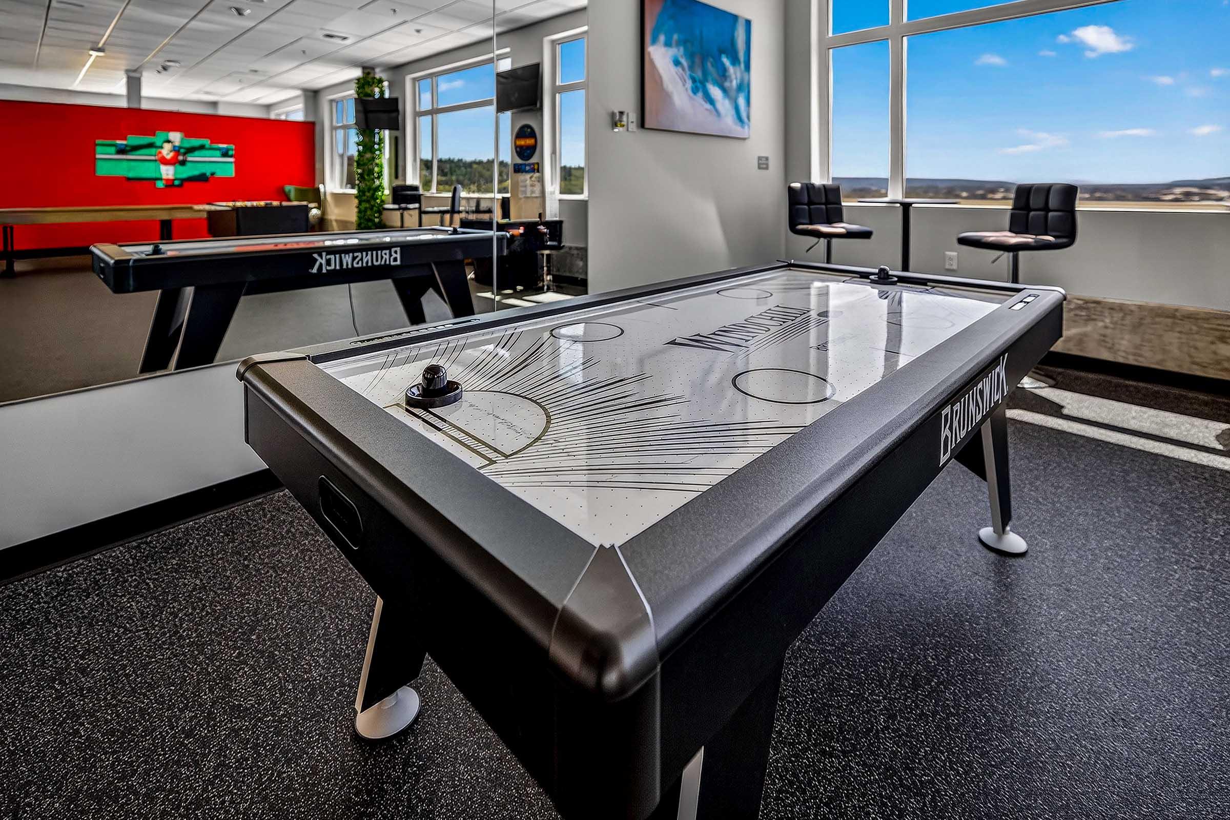 A modern office game room featuring a sleek air hockey table in the foreground. The background shows a vibrant red wall with a colorful artwork and a lounge area with chairs. Large windows provide a scenic view, filling the space with natural light. The flooring is dark, complementing the contemporary decor.