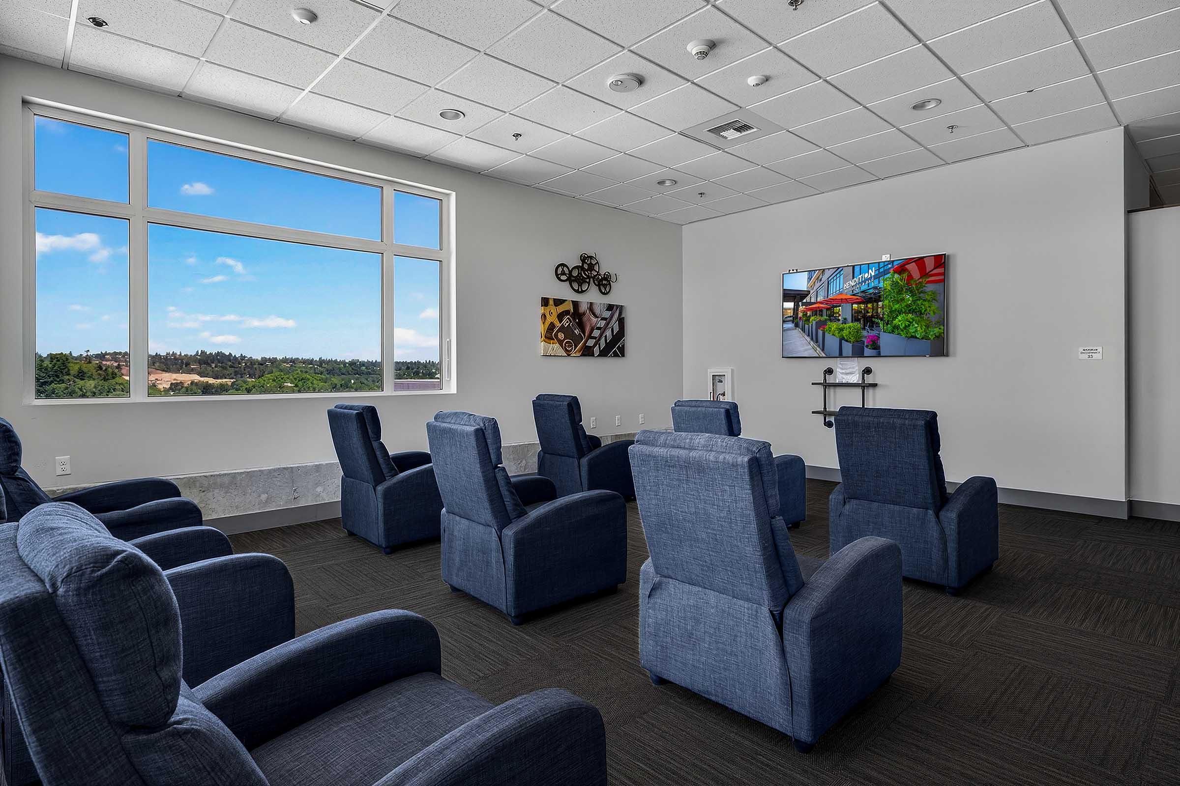 A well-lit lounge area featuring several blue recliner chairs arranged facing a wall-mounted TV. Large windows provide a view of a green landscape and blue sky. The walls are decorated with modern artwork, creating a comfortable space for relaxation or socializing.