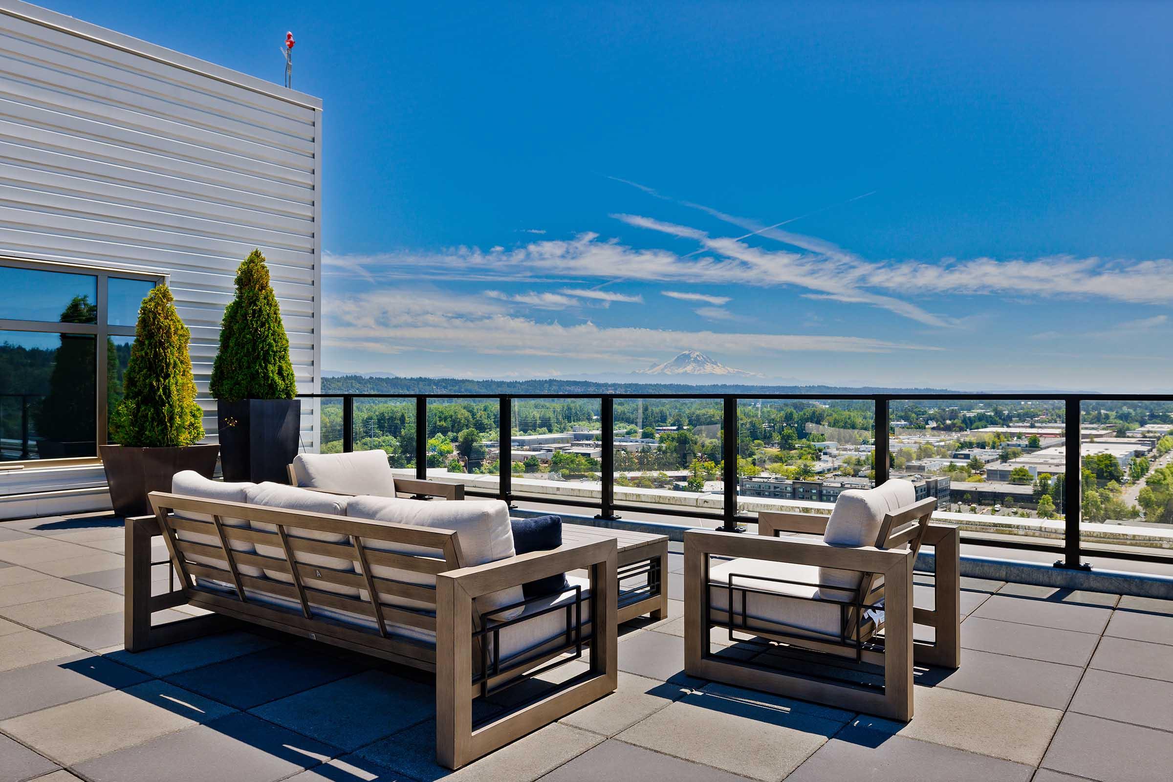 A rooftop patio featuring modern outdoor furniture, including a stylish couch and chairs, overlooking a scenic landscape with mountains in the distance. The sky is bright and blue, with a few clouds, and greenery can be seen in the vicinity, creating a serene and inviting atmosphere.