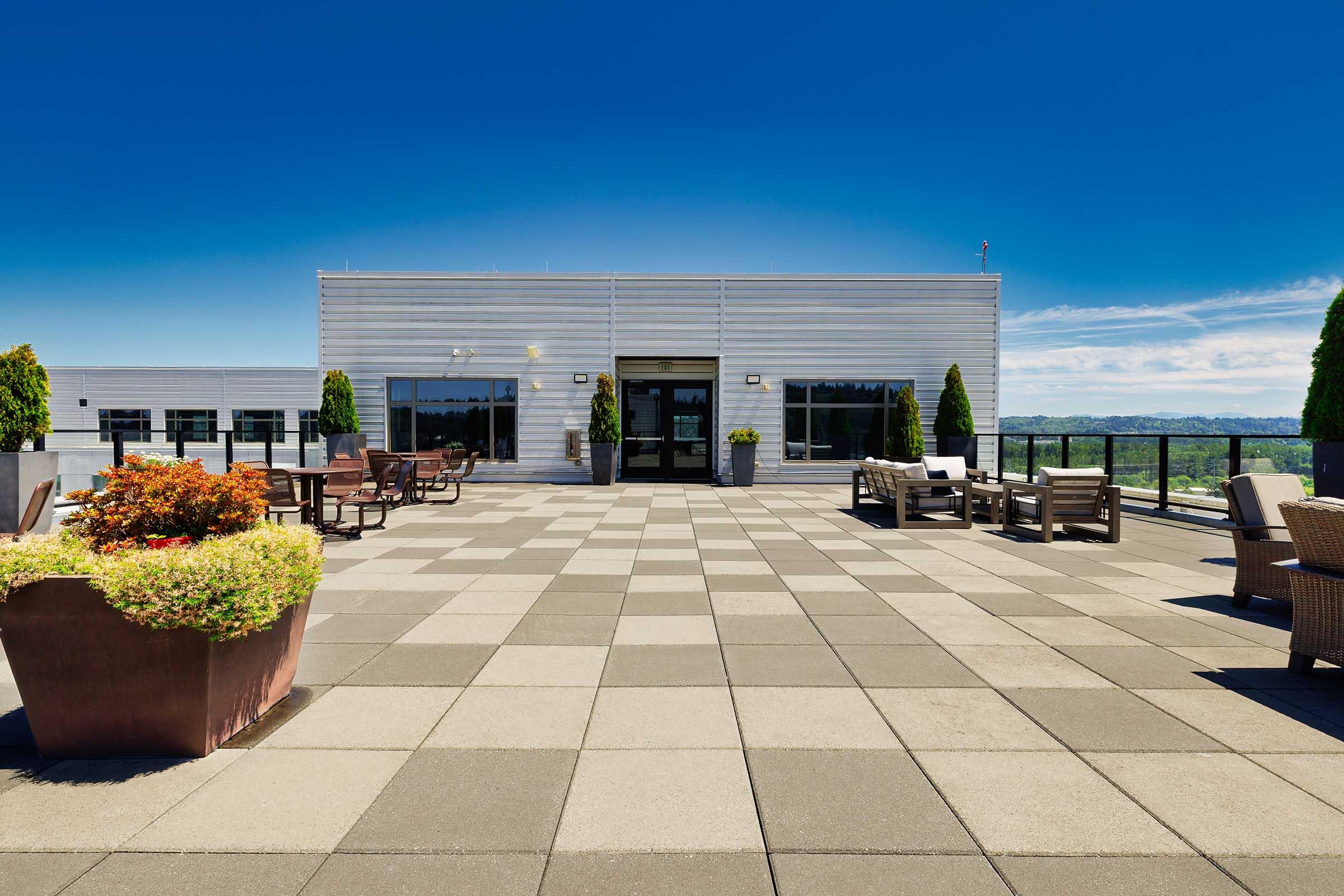 A spacious rooftop terrace featuring a checkered tiled floor, outdoor seating arrangements including chairs and tables, and decorative planters. The background showcases a clear blue sky and distant hills, creating a serene atmosphere for relaxation or gatherings.