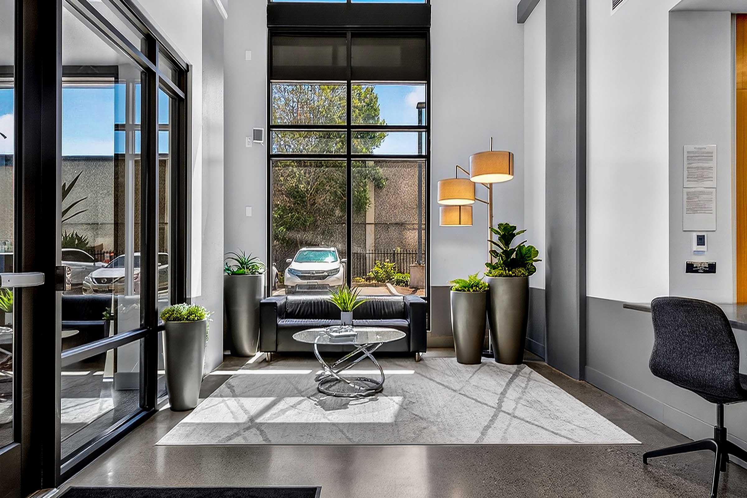 A modern lobby featuring large windows allowing natural light, a sleek black sofa, decorative plants, and stylish lighting. The space has a minimalist design with a round coffee table and a gray rug, creating a welcoming atmosphere. Outside, a car is visible through the glass doors.