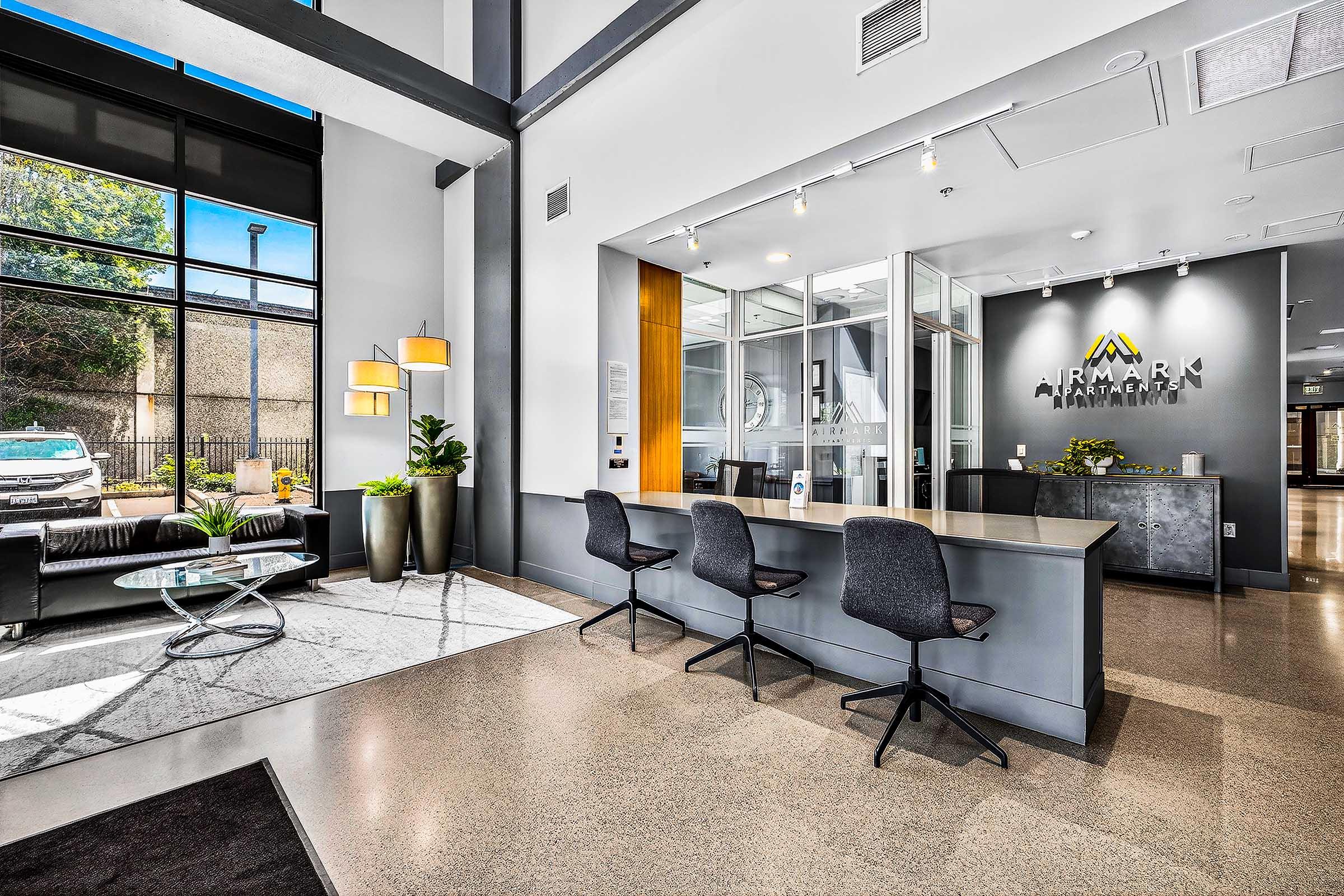 Modern office lobby featuring a sleek reception desk, stylish seating area with a coffee table, and large windows allowing natural light. Elegant decor includes potted plants and contemporary lighting fixtures, creating a welcoming atmosphere. A parked car is visible outside through the glass.