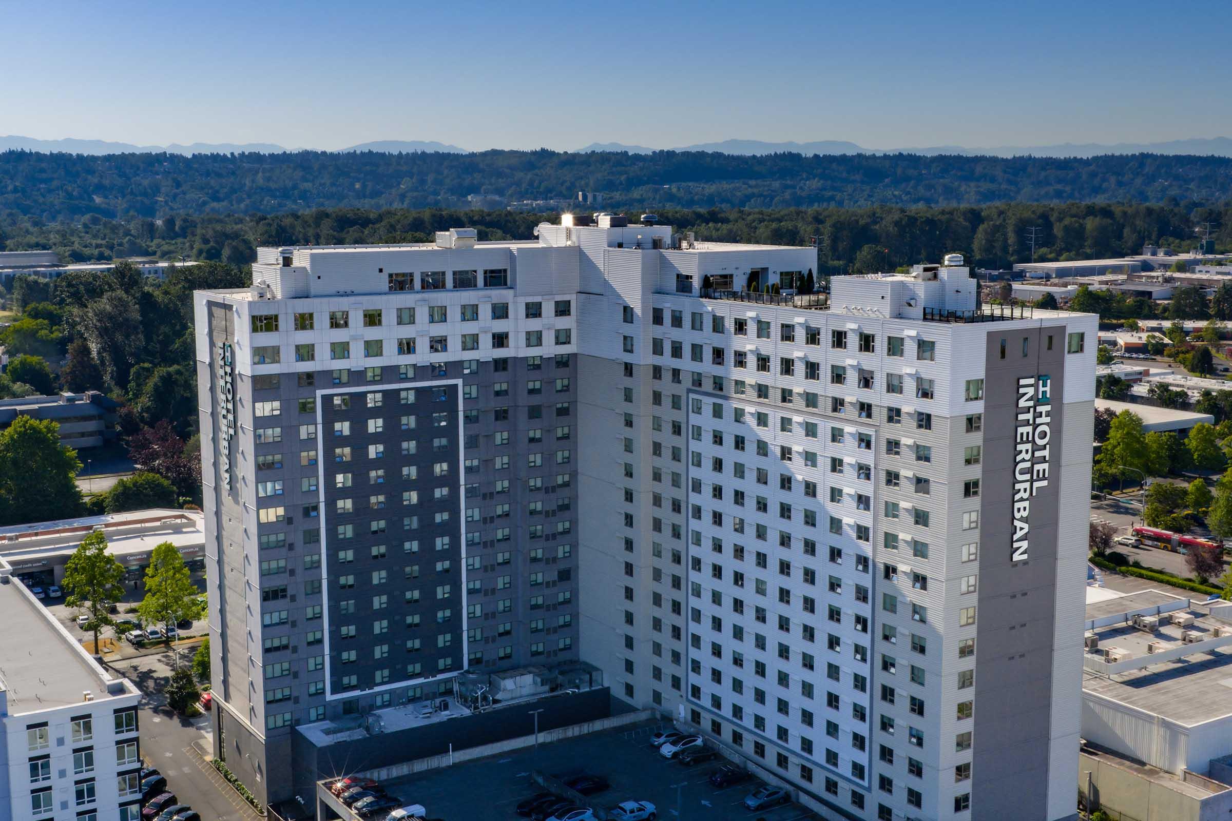 Aerial view of the InterContinental Hotel, showcasing its modern architecture. The hotel features a tall, multi-story building with a mix of gray and white panels. Surrounding green trees and a clear blue sky create a vibrant backdrop, highlighting the hotel’s urban location. Parking area visible in the foreground.