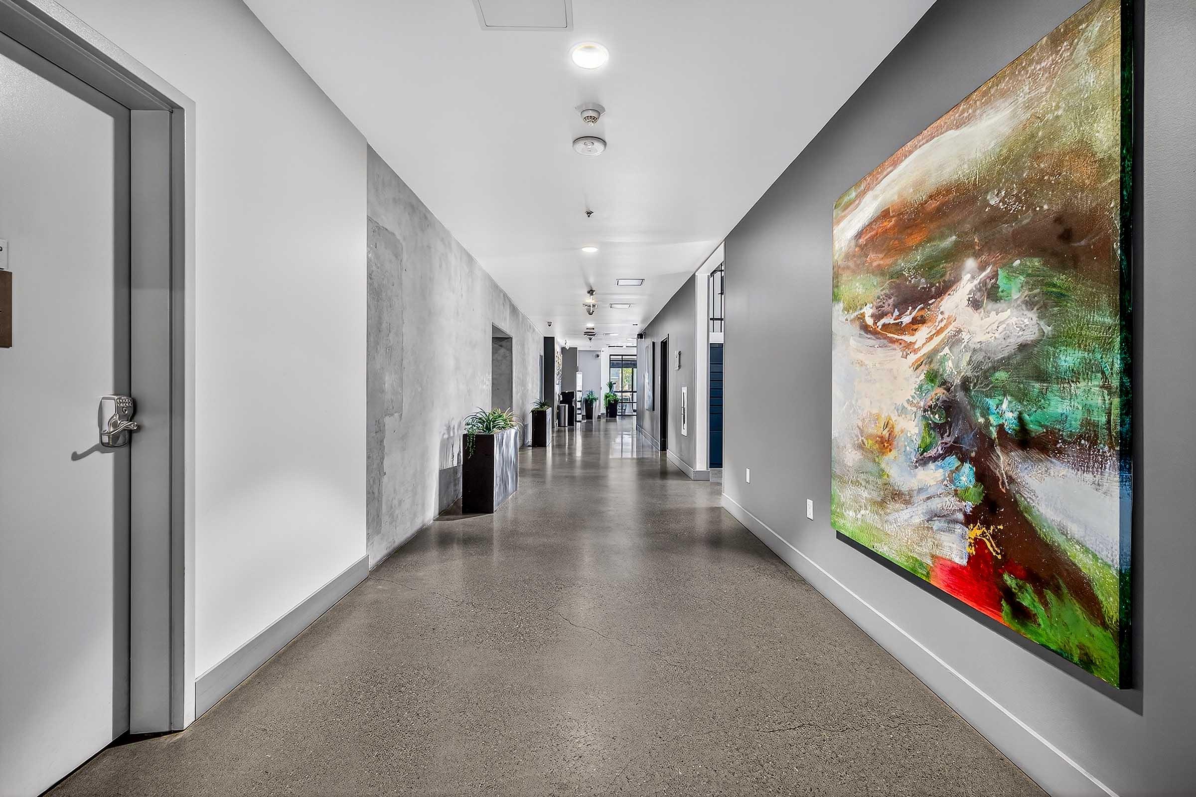 A well-lit corridor featuring a polished concrete floor. On one side, there is a colorful abstract painting mounted on a gray wall. Potted plants are positioned along the hallway, leading towards a doorway at the far end. The overall aesthetic is modern and inviting.