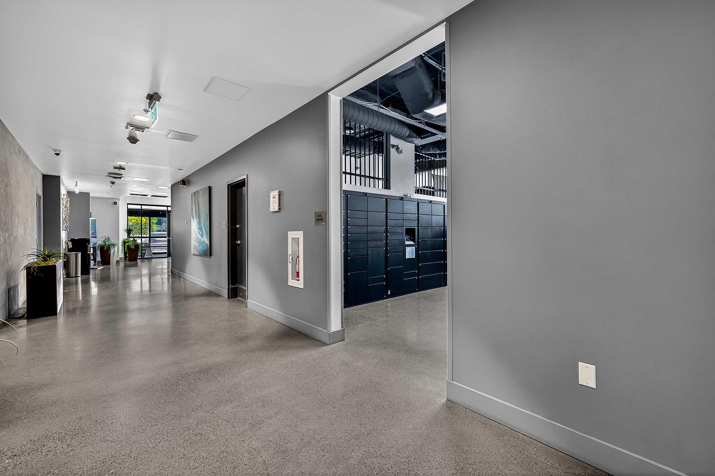 A modern hallway featuring a sleek design with gray walls and polished concrete flooring. On one side, there is a vibrant artwork, and on the other, a row of black lockers. Natural light is coming through large windows at the far end, creating an inviting atmosphere.