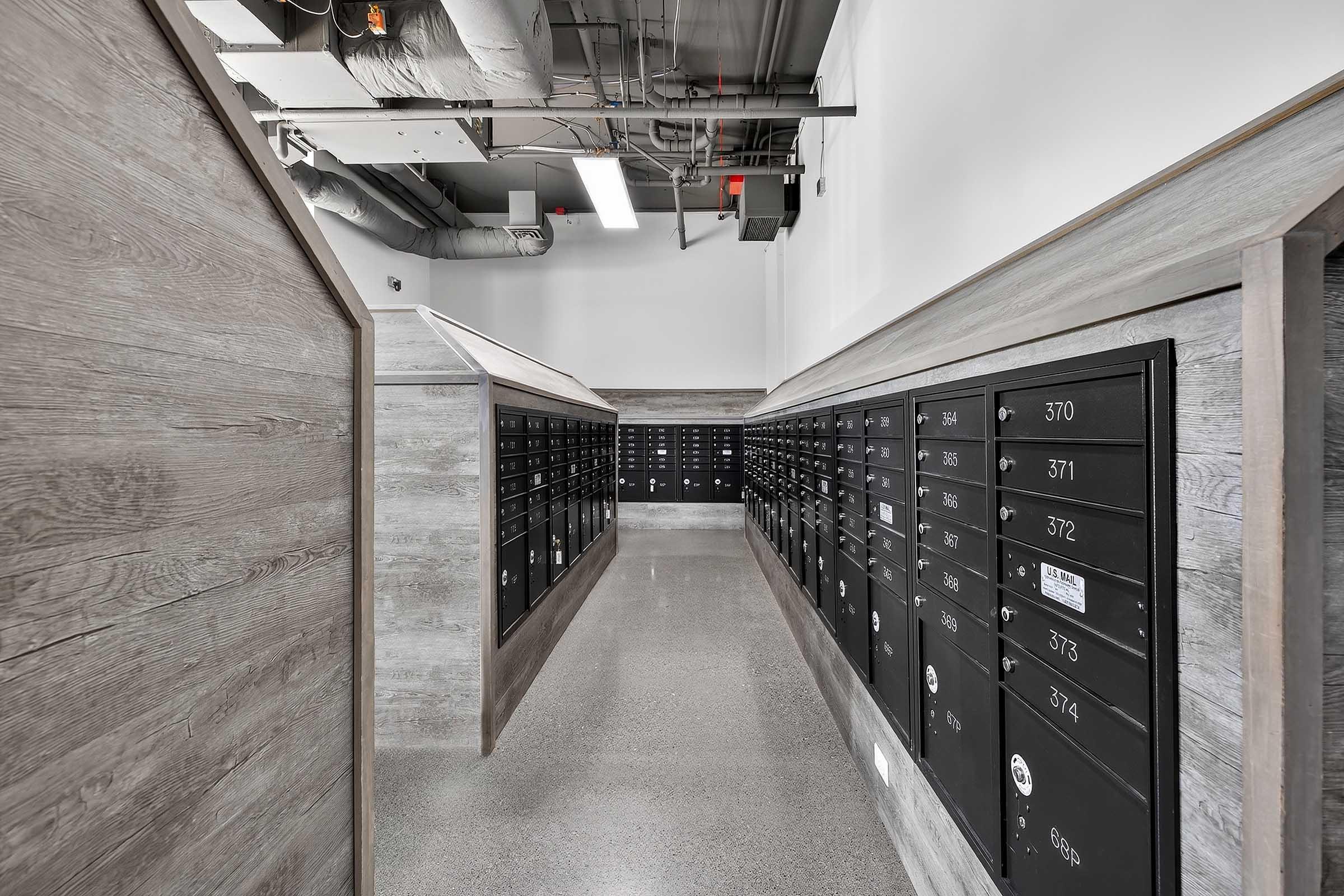 Interior view of a modern mailroom featuring rows of black mail lockers against a minimalist design with wooden accents. The space is well-lit and has an industrial feel, with exposed ductwork and smooth flooring.