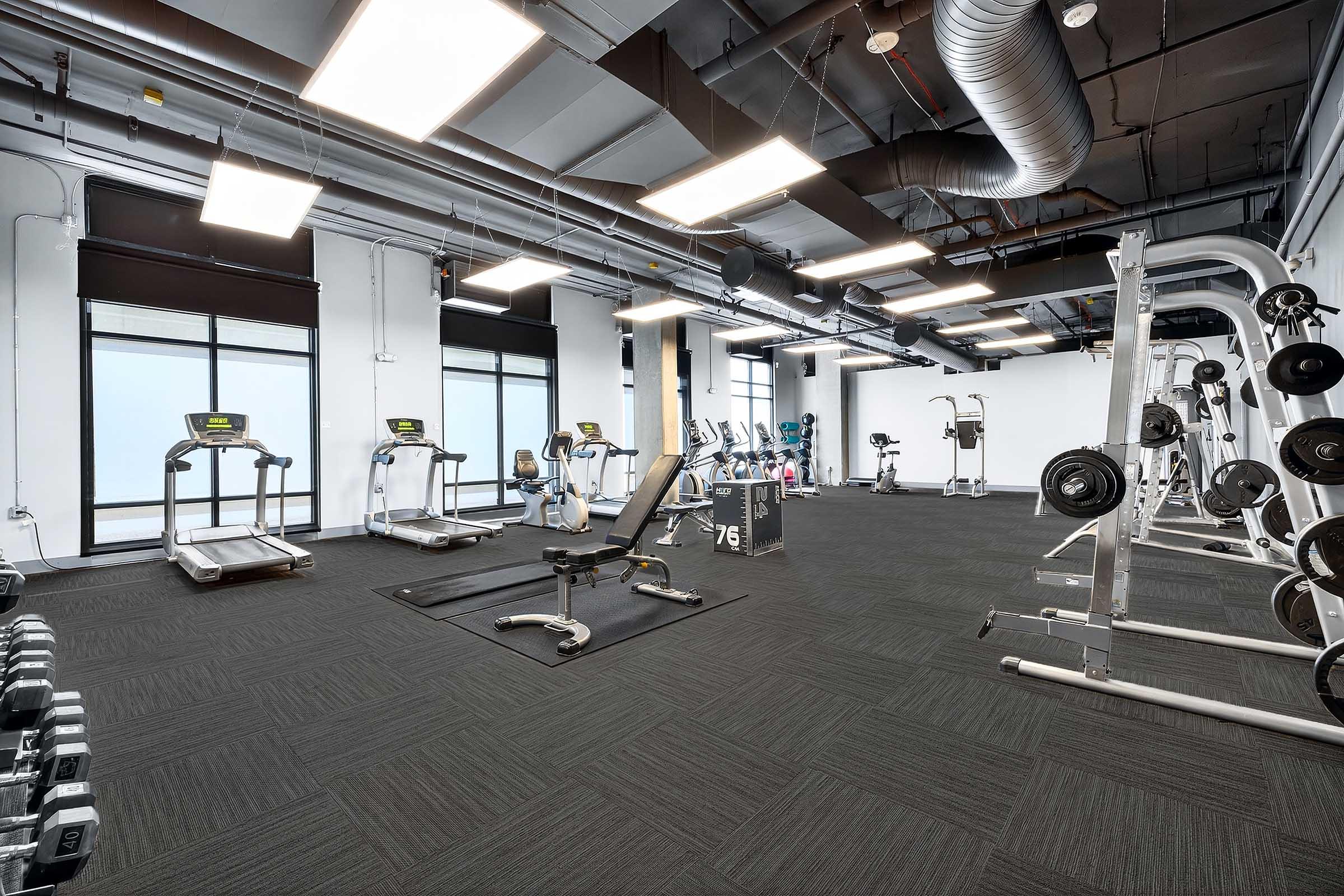 A spacious, modern gym featuring several treadmills, weightlifting equipment, and free weights. The room has large windows allowing natural light in, a clean floor with a dark carpet, and high ceilings with industrial-style ventilation.
