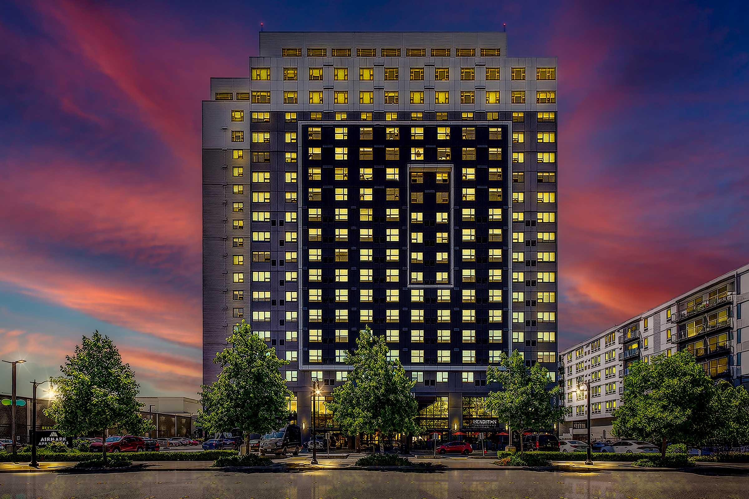 A modern hotel building illuminated at dusk, showcasing large windows with warm lights. The facade features a geometric design with a dark exterior. Lush trees frame the entrance, and nearby low-rise buildings are visible. The sky is a blend of purple and orange hues, creating a vibrant sunset backdrop.