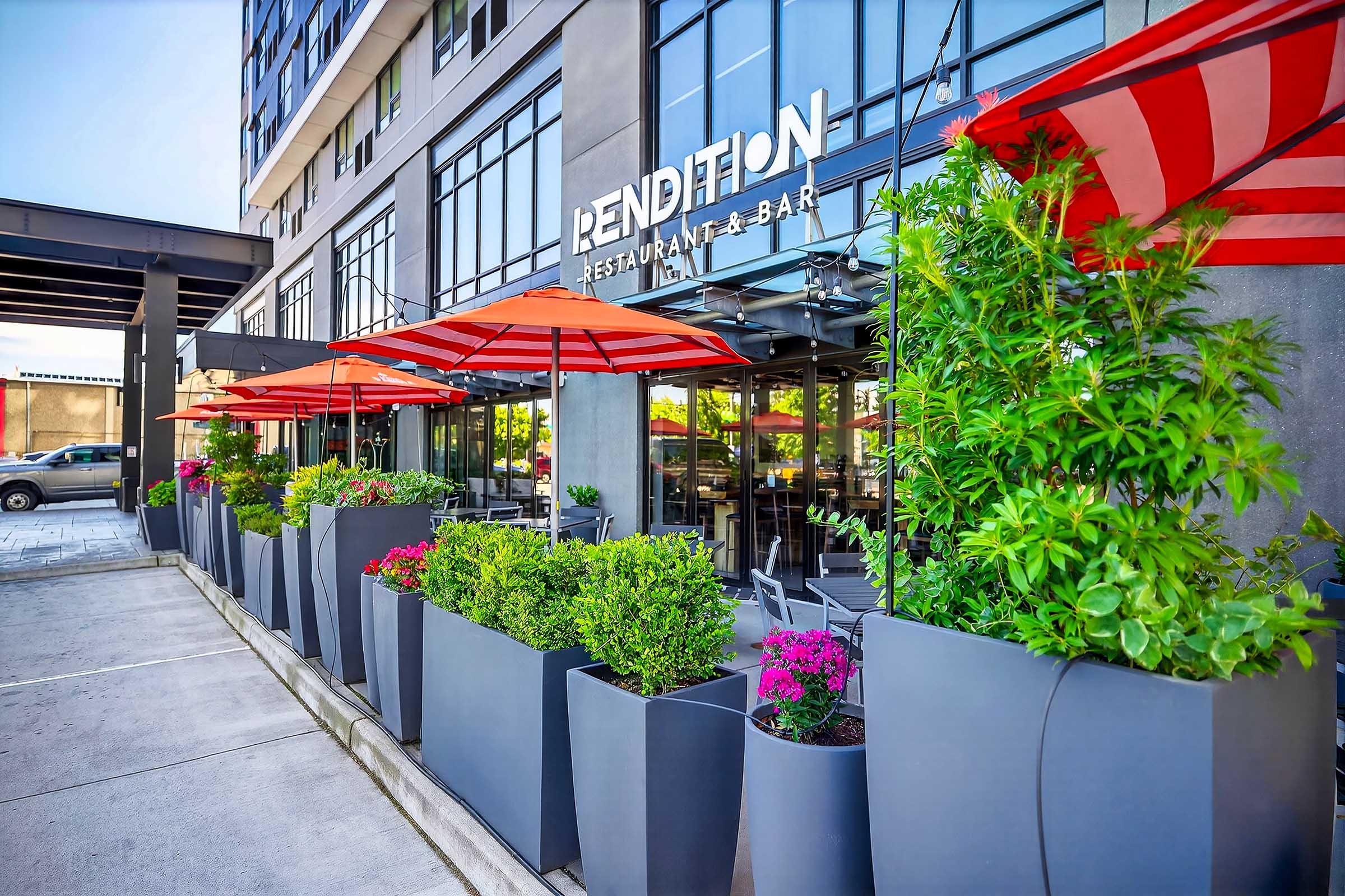 Exterior view of Rendition Restaurant & Bar featuring large planters with colorful flowers and greenery. Red and white umbrellas provide shade for outdoor seating areas. The building has large glass windows and a modern architectural design. The setting is bright and inviting, suggesting a lively dining atmosphere.