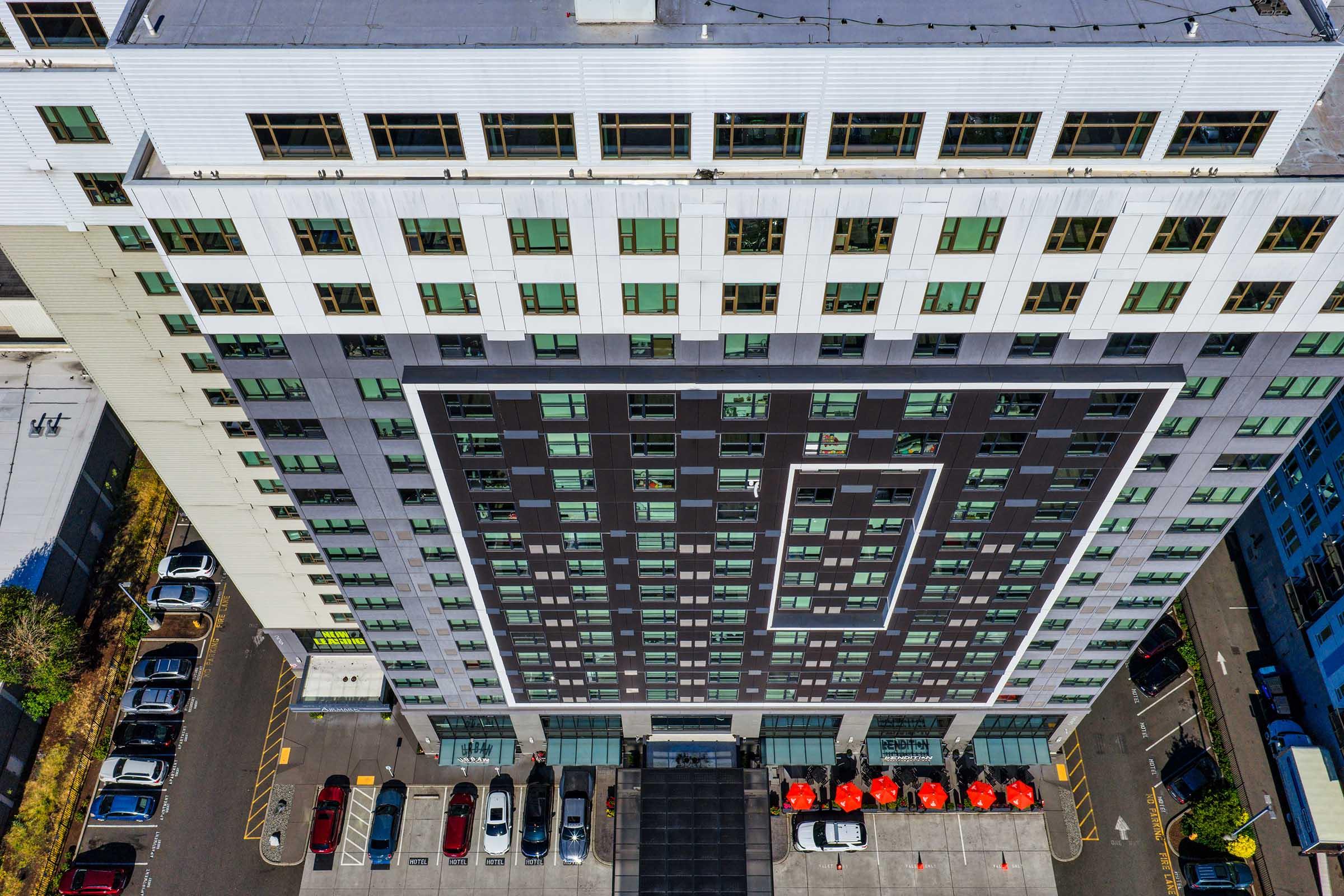 Aerial view of a modern multi-story building featuring a mix of dark and light-colored exterior panels, large windows, and a flat roof. The ground level includes a parking area with several vehicles and outdoor seating under orange umbrellas. Surrounding greenery adds to the urban environment.
