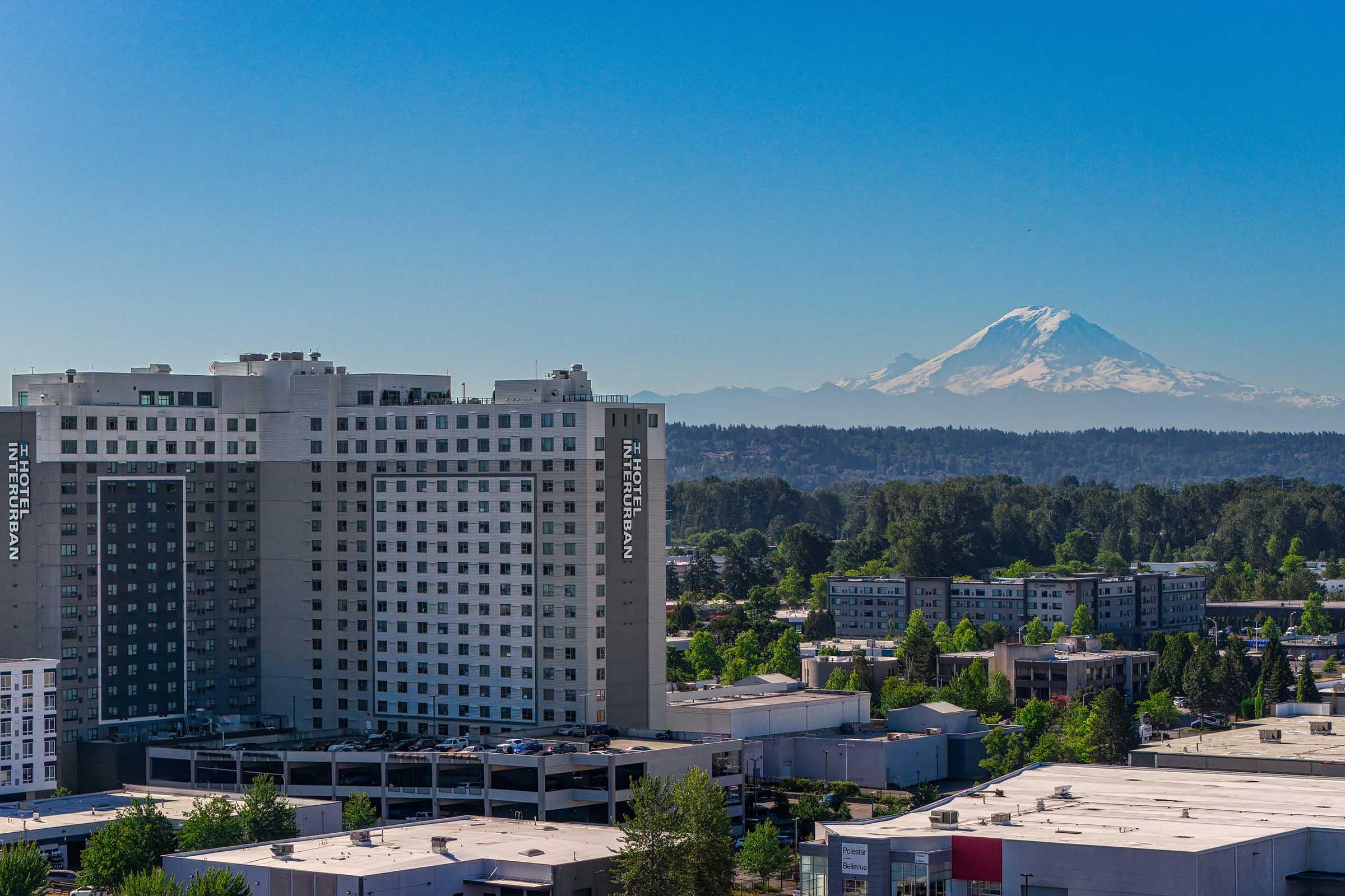 A panoramic view featuring a modern high-rise building in the foreground, with a clear blue sky above. In the distance, a snow-capped mountain is visible, surrounded by green trees and urban structures, creating a contrasting landscape of natural beauty and urban development.