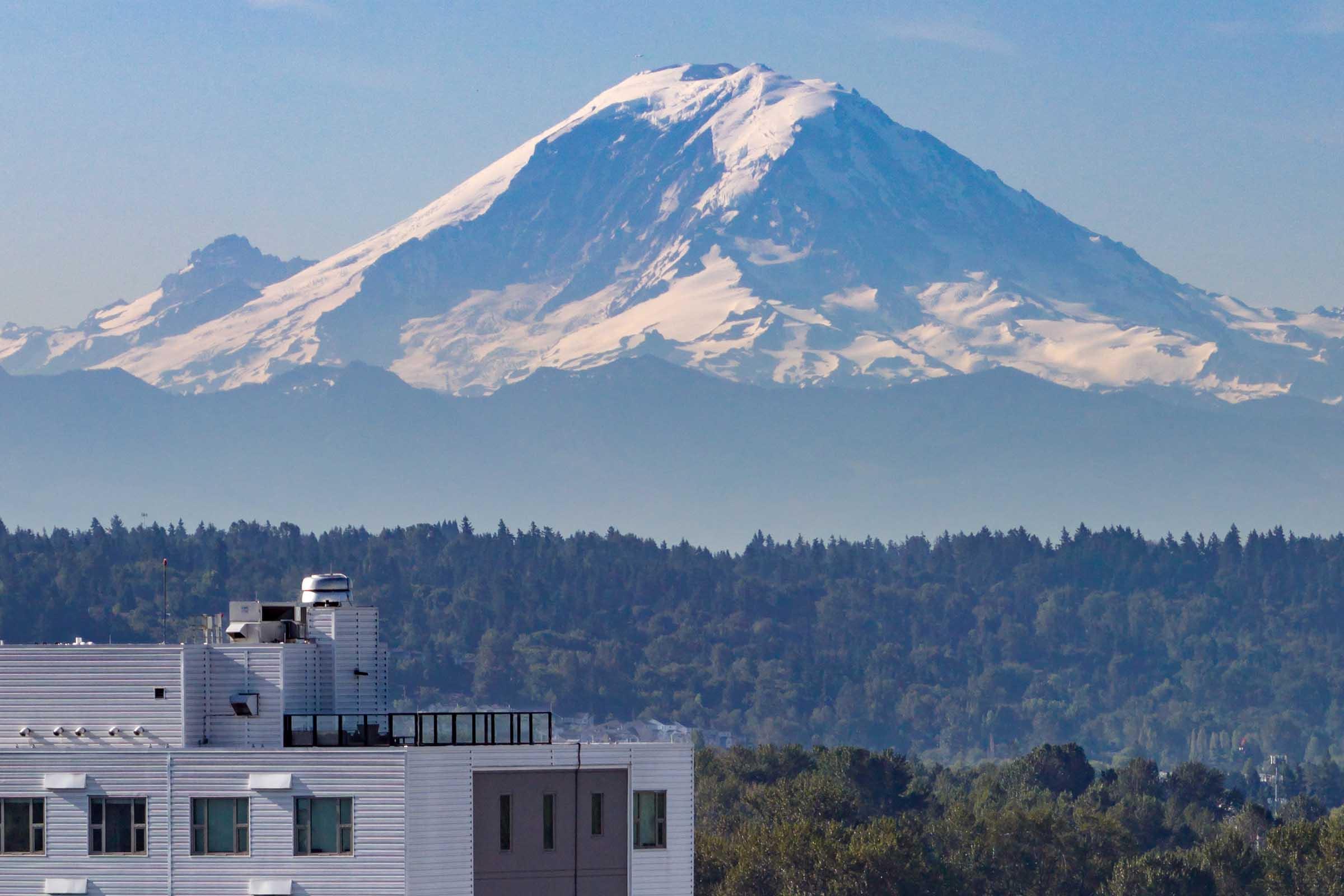 A snow-capped mountain rises majestically in the background, with lush green forests at its base. In the foreground, a modern building with a rooftop structure is visible, framed by the natural landscape. Clear blue skies enhance the serene and picturesque scenery.