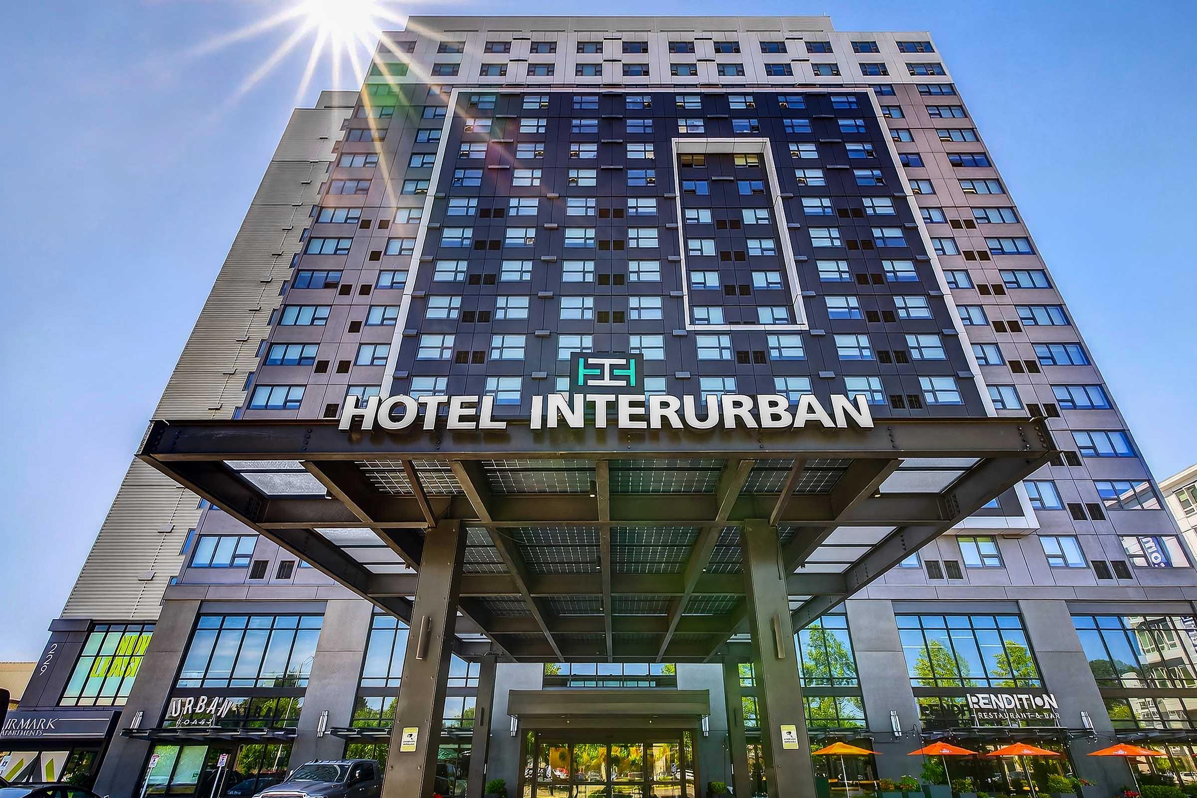 A modern building facade of Hotel Interurban featuring a large entrance with a canopy. The hotel has a sleek design with a combination of grey and blue panels. Bright sunlight is shining from the top left corner of the image, highlighting the structure against a clear blue sky. Outdoor seating areas are visible in front.