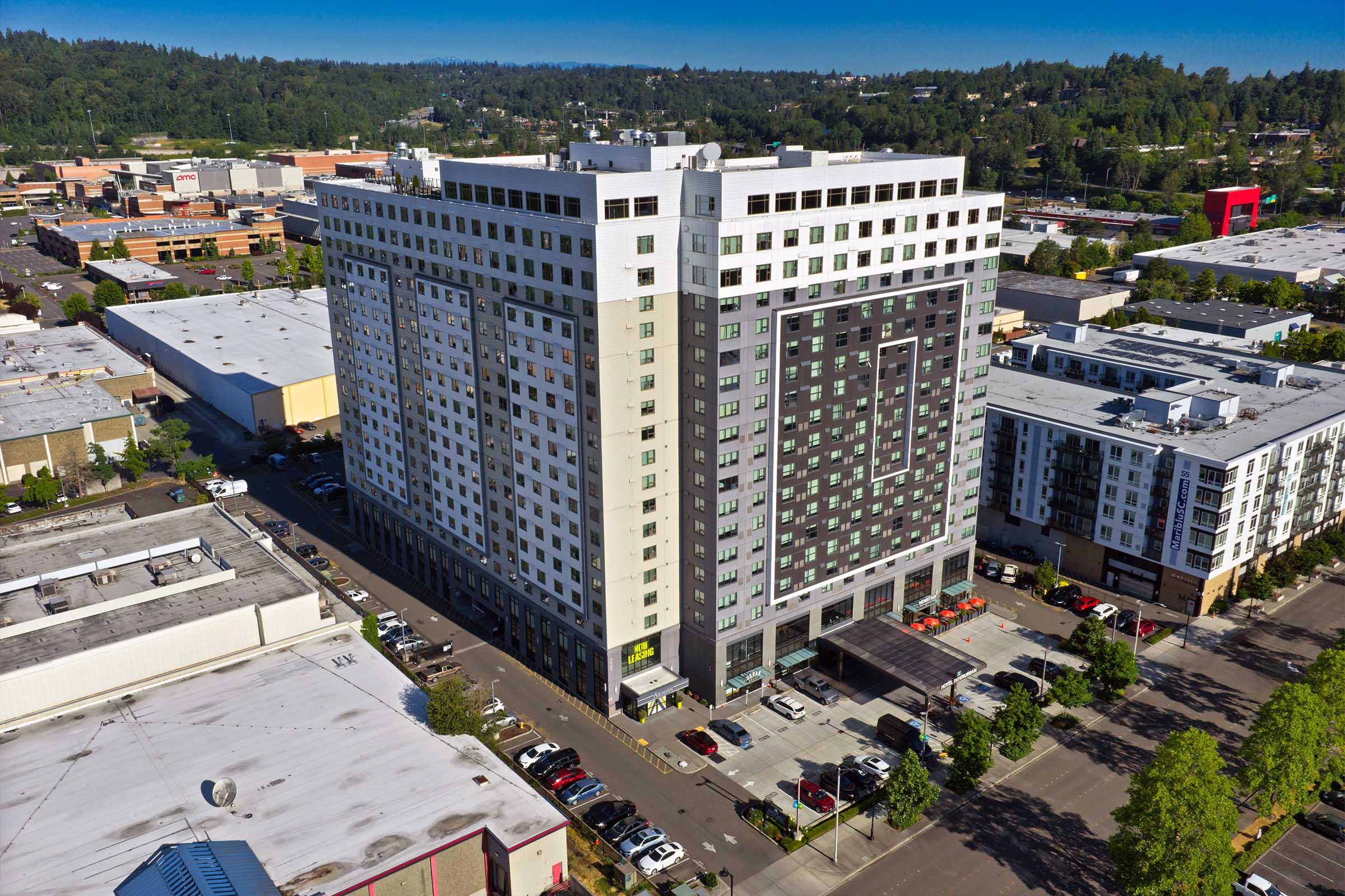 Aerial view of a modern multi-story building featuring a mix of light and dark gray facade. The structure has large windows and is surrounded by parking spaces and nearby retail developments. Green trees and a clear blue sky are visible in the background, indicating a vibrant urban environment.