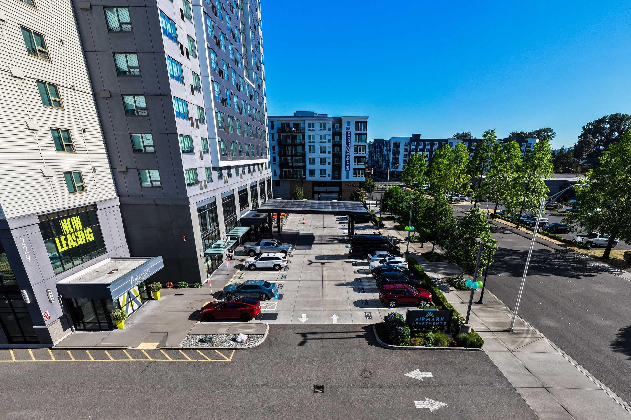 Aerial view of a modern residential building with "Now Leasing" signage. Below, a parking lot features several cars and designated parking spaces, surrounded by trees. Nearby, additional multi-story apartment buildings are visible under a clear blue sky.